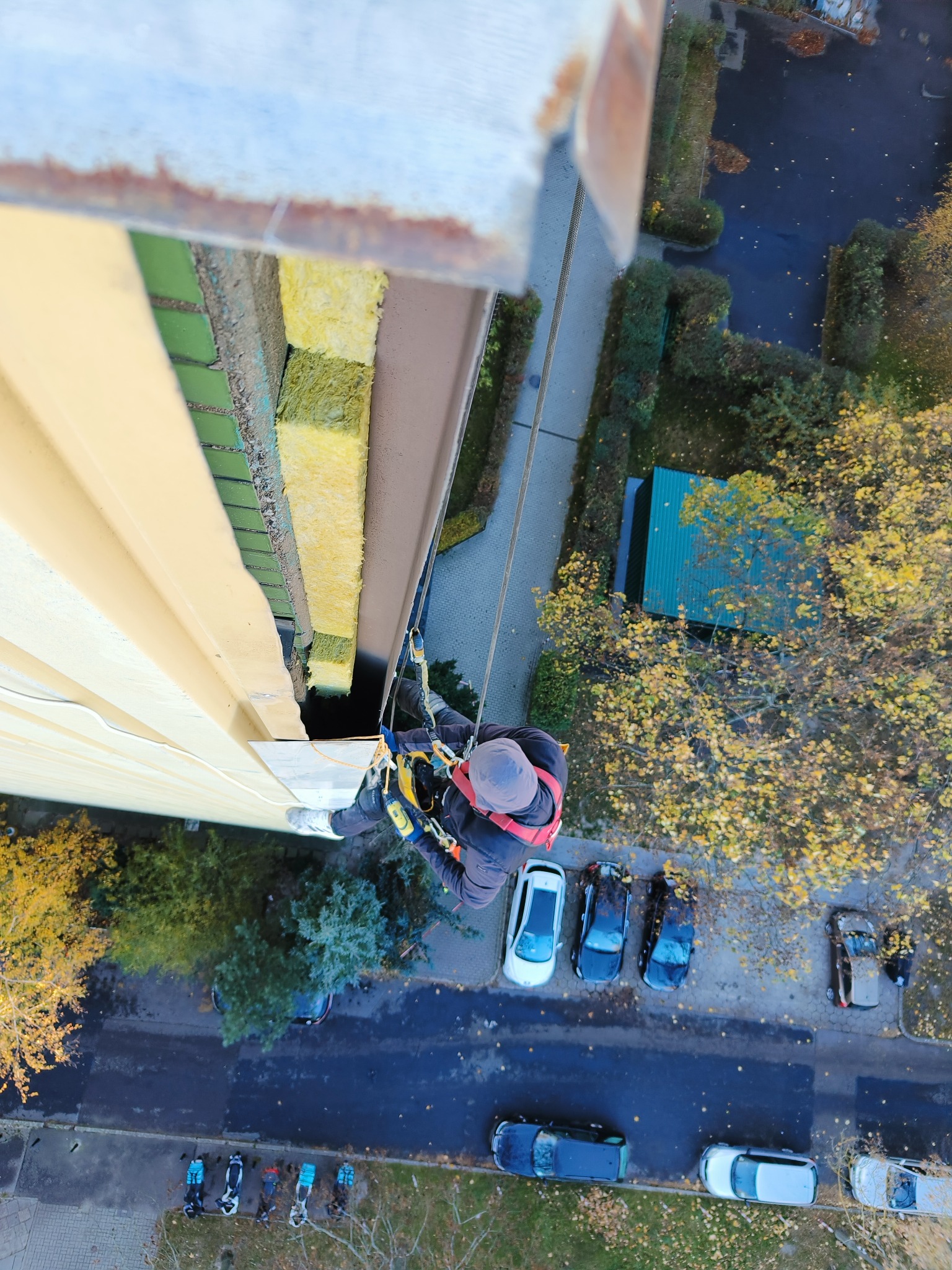 Pracownik w uprzęży, zawieszony na linie, wykonuje prace na wysokości na elewacji budynku. Widok z góry na pracownika, fragment elewacji z widoczną izolacją, ulicę z zaparkowanymi samochodami...