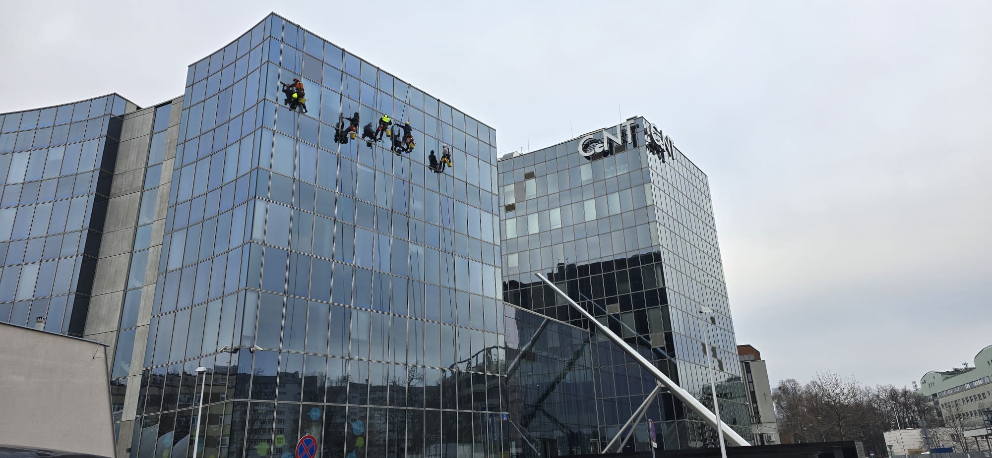 Grupa alpinistów przemysłowych w uprzężach, czyszczących szklaną fasadę biurowca w pochmurny dzień. Widoczne wiadra i akcesoria do mycia szyb.