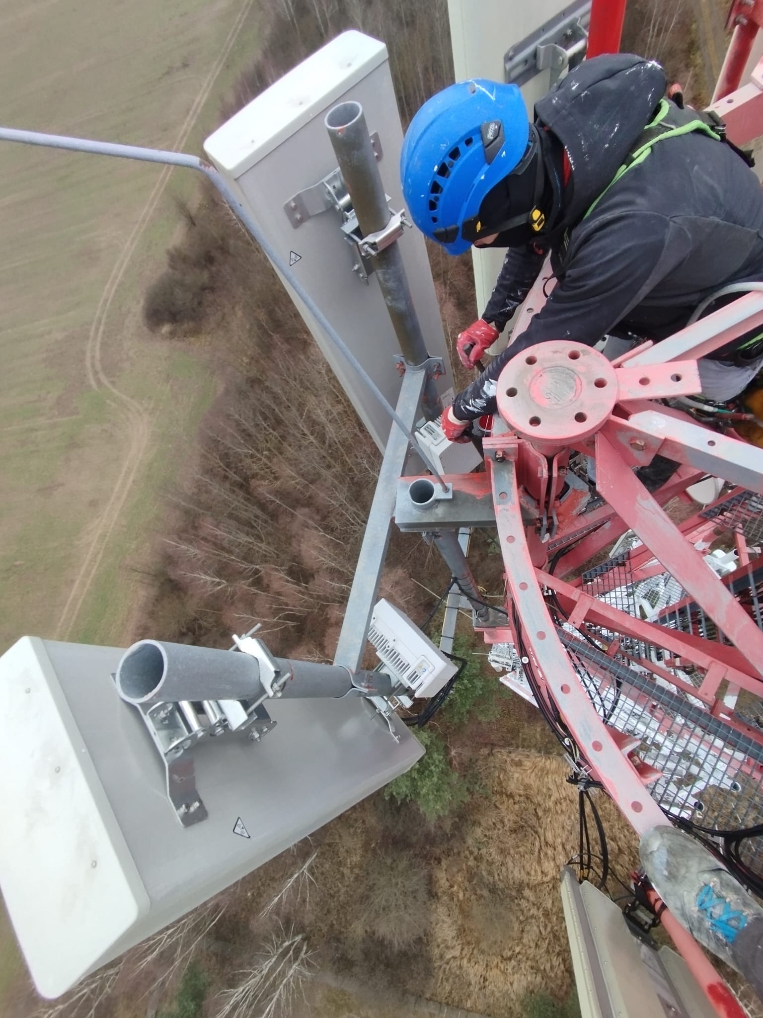 Pracownik w niebieskim kasku Petzl, zabezpieczony linami, montuje antenę na czerwonej konstrukcji wieży telekomunikacyjnej; widok z góry na las i pole.