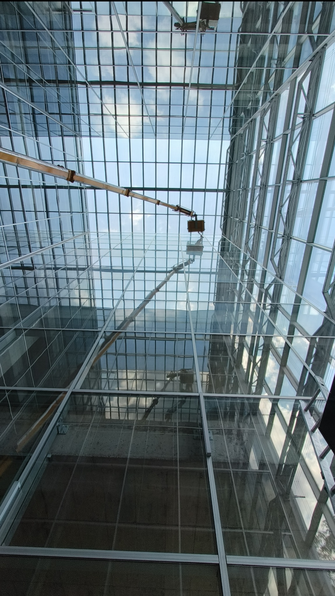 Widok z dołu na szklany dach budynku podczas czyszczenia za pomocą podnośnika koszowego. Refleksy nieba w czyszczonych oknach.