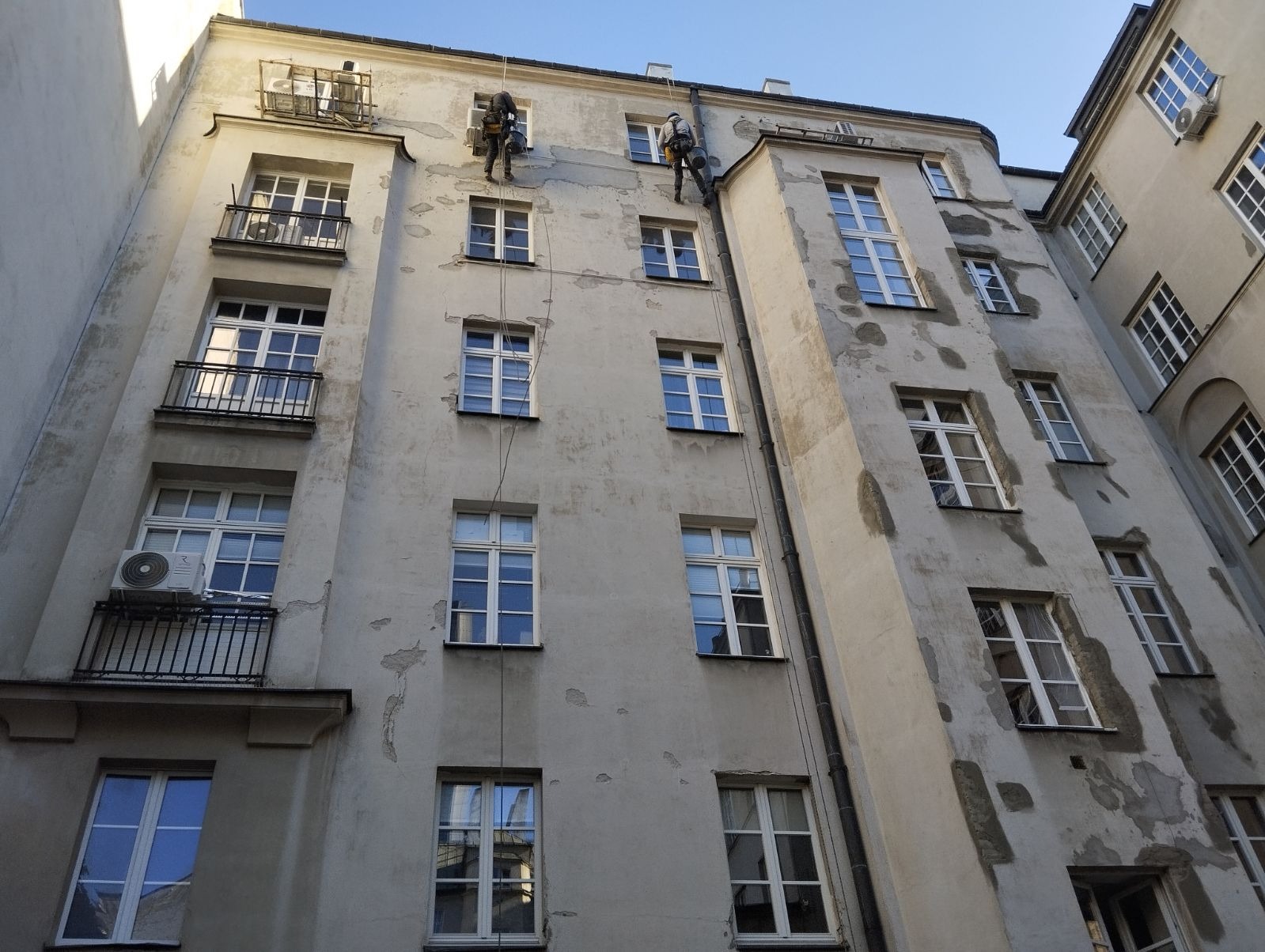 Dwóch alpinistów na linach podczas renowacji elewacji kamienicy z balkonami i klimatyzacją, widok z dołu na tle błękitnego nieba.