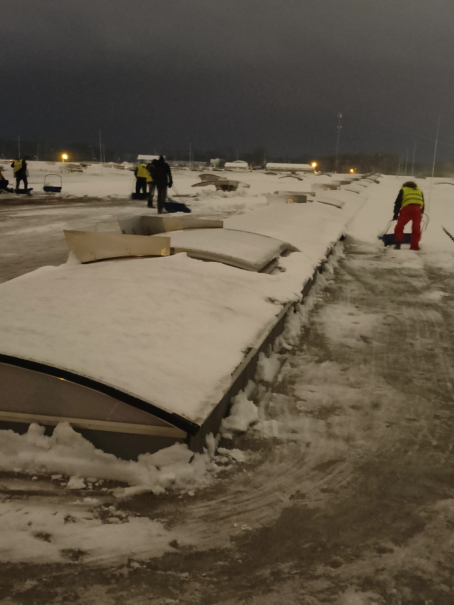 Pracownicy w odblaskowych kamizelkach usuwają śnieg z dachu budynku o zmierzchu, używając łopat i odśnieżarek ręcznych.