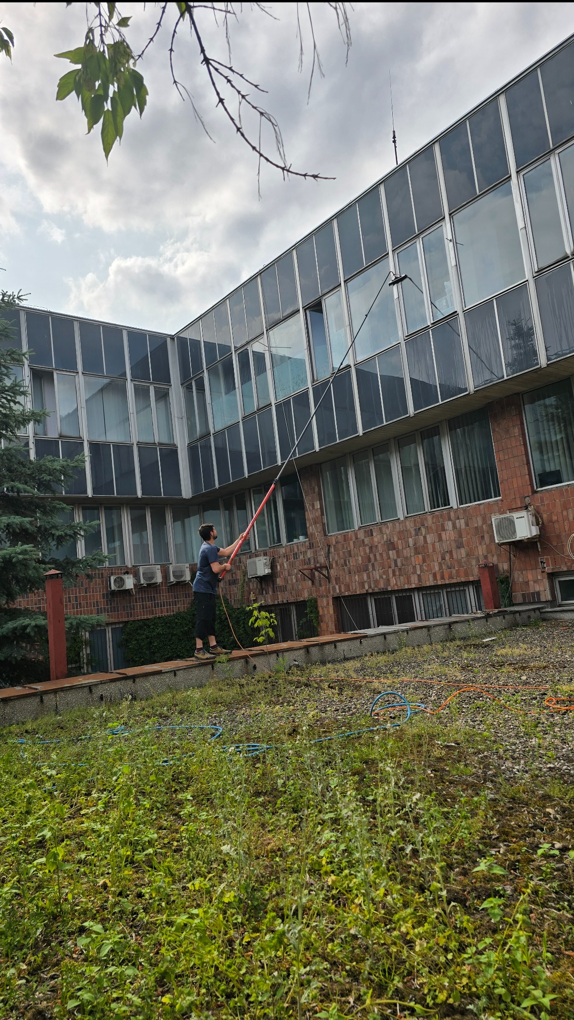 Mężczyzna myje okna budynku biurowego za pomocą długiej lancy teleskopowej, stojąc na podwyższeniu wśród zieleni. Widoczne okna z refleksami i ceglana elewacja.