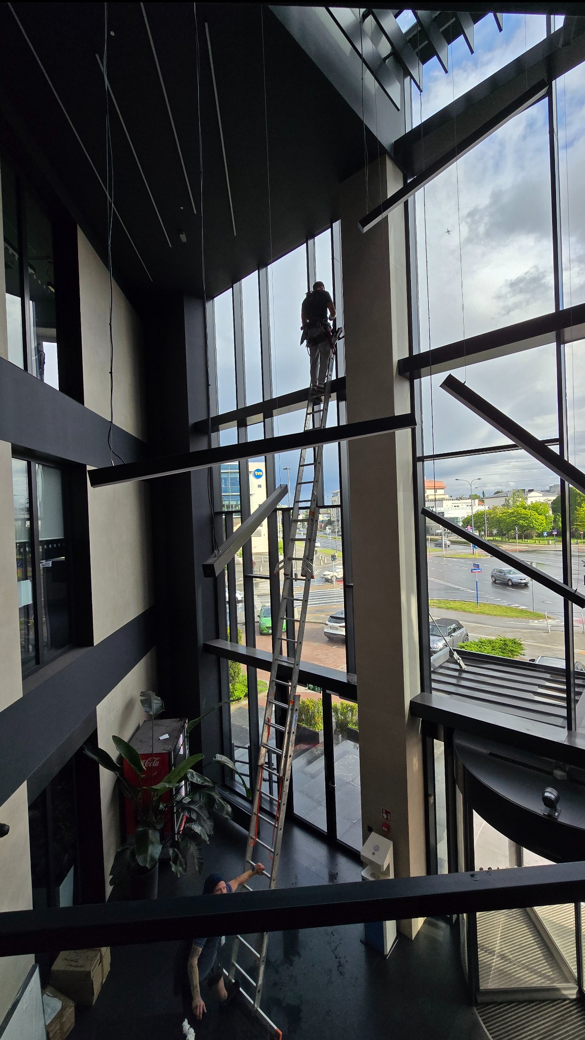 Pracownik na drabinie, w uprzęży, czyści wysokie okna w nowoczesnym biurowcu. Widok z góry, na zewnątrz widoczny ruch uliczny i budynki.