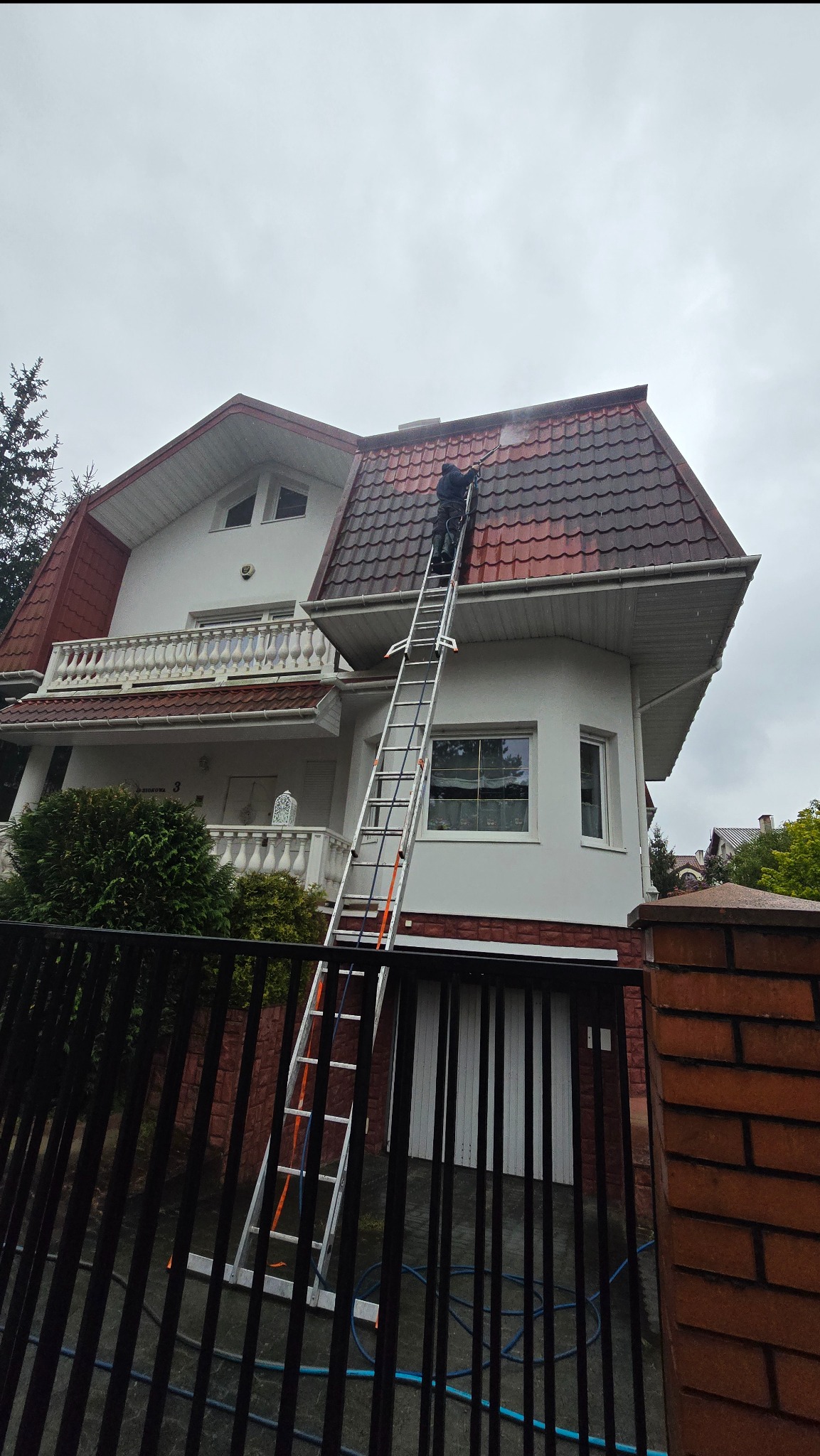 Pracownik na drabinie czyści ciśnieniowo dach dwupiętrowego domu z czerwonej dachówki. Widoczne ogrodzenie posesji i fragment elewacji. Pochmurne niebo.
