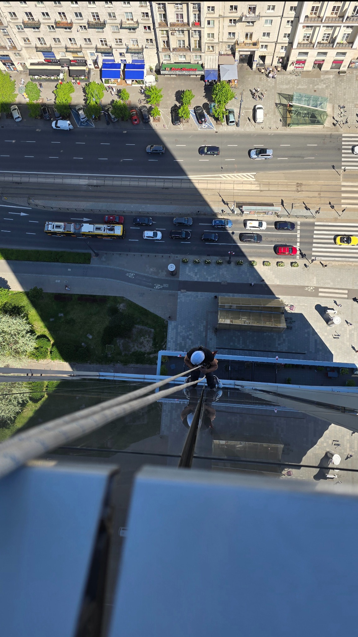 Pracownik na wysokości myje szklaną elewację biurowca, widok z góry na ulicę z ruchem miejskim. Bezpieczeństwo zapewniają liny i kask.