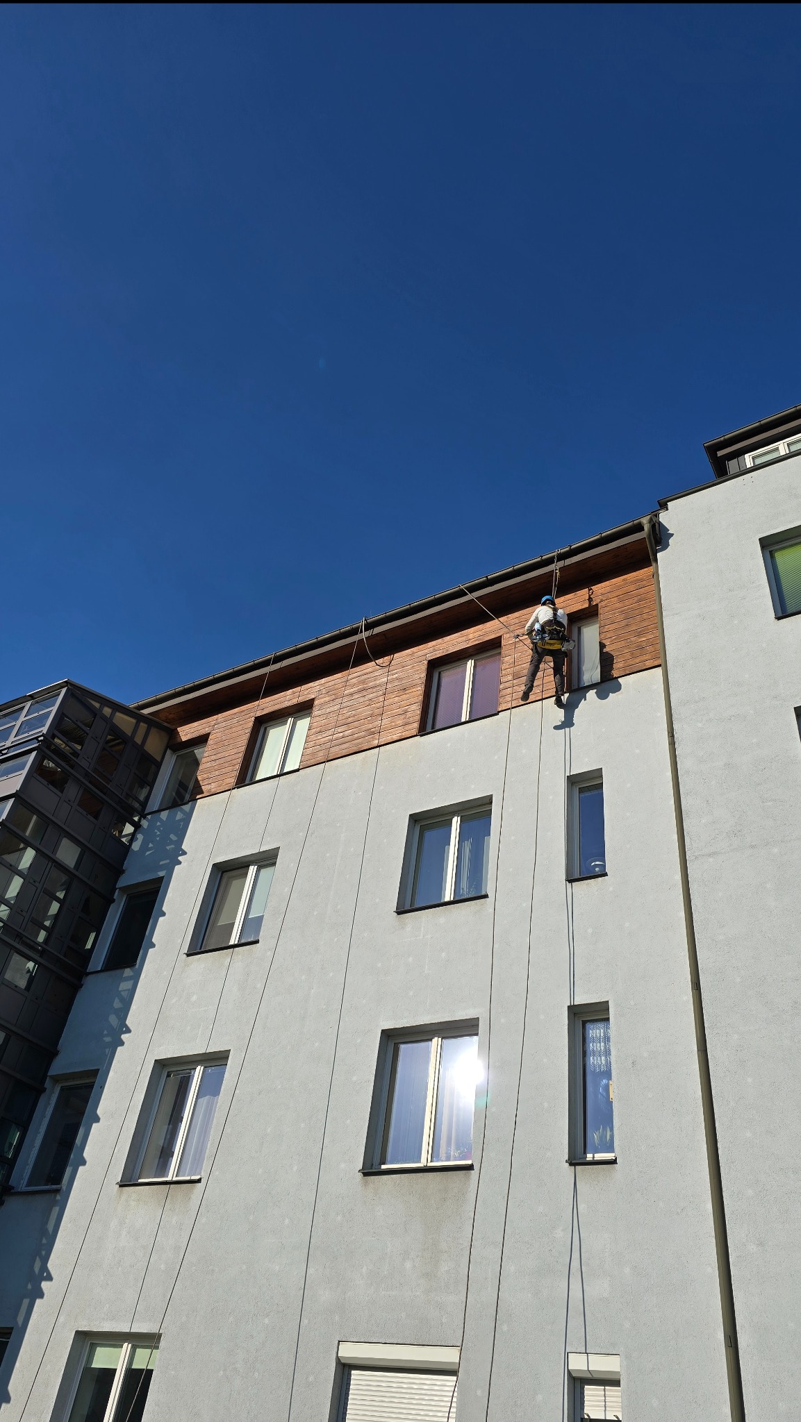 Pracownik na linie, w uprzęży, czyści okna na wysokości w budynku z szarą elewacją i drewnianą górną częścią. Widok z dołu, błękitne niebo.