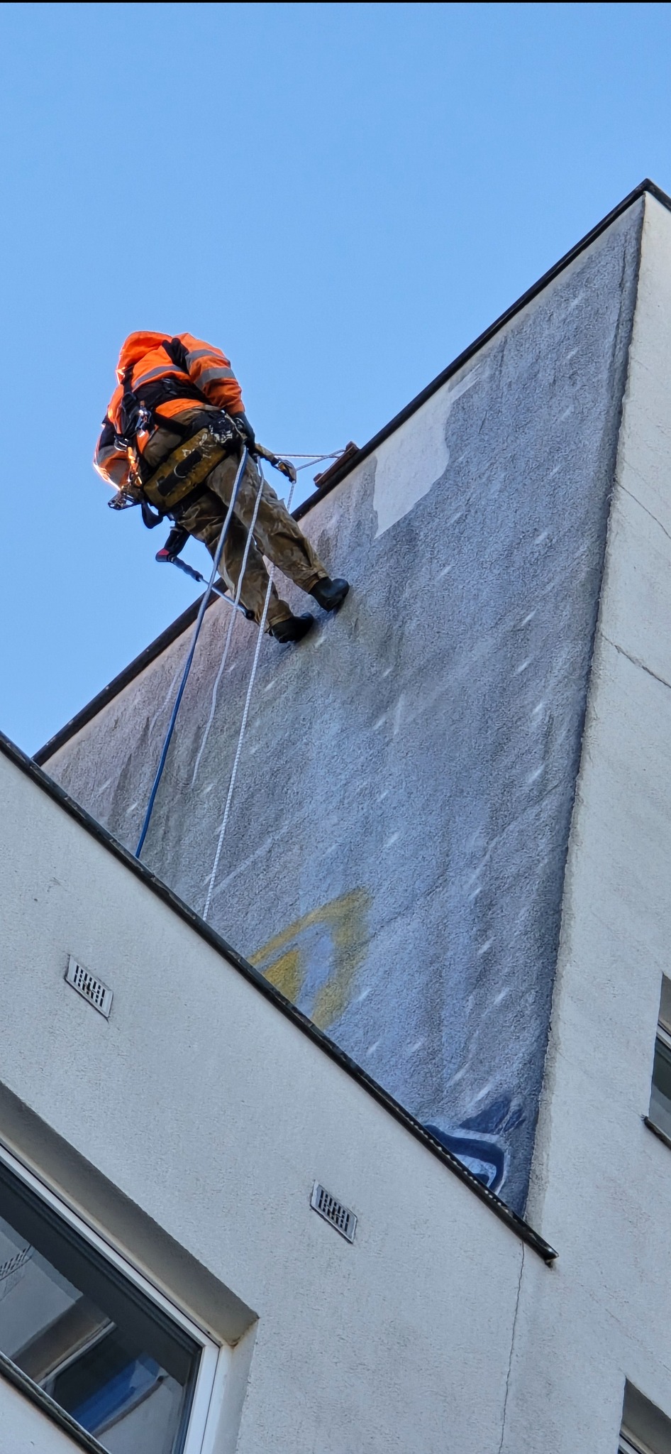 Pracownik w uprzęży, na linie, maluje elewację budynku. Widoczne fragmenty pomalowanej ściany i resztki graffiti. Ujęcie z dołu, na tle błękitnego nieba.