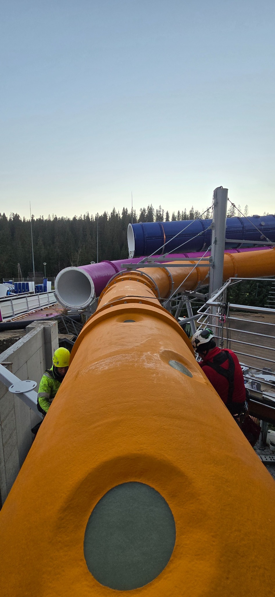 Dwóch pracowników w kaskach i uprzężach pracuje przy montażu pomarańczowej rury z włókna szklanego na wysokości, elementu parku wodnego z kolorowymi zjeżdżalniami w tle.