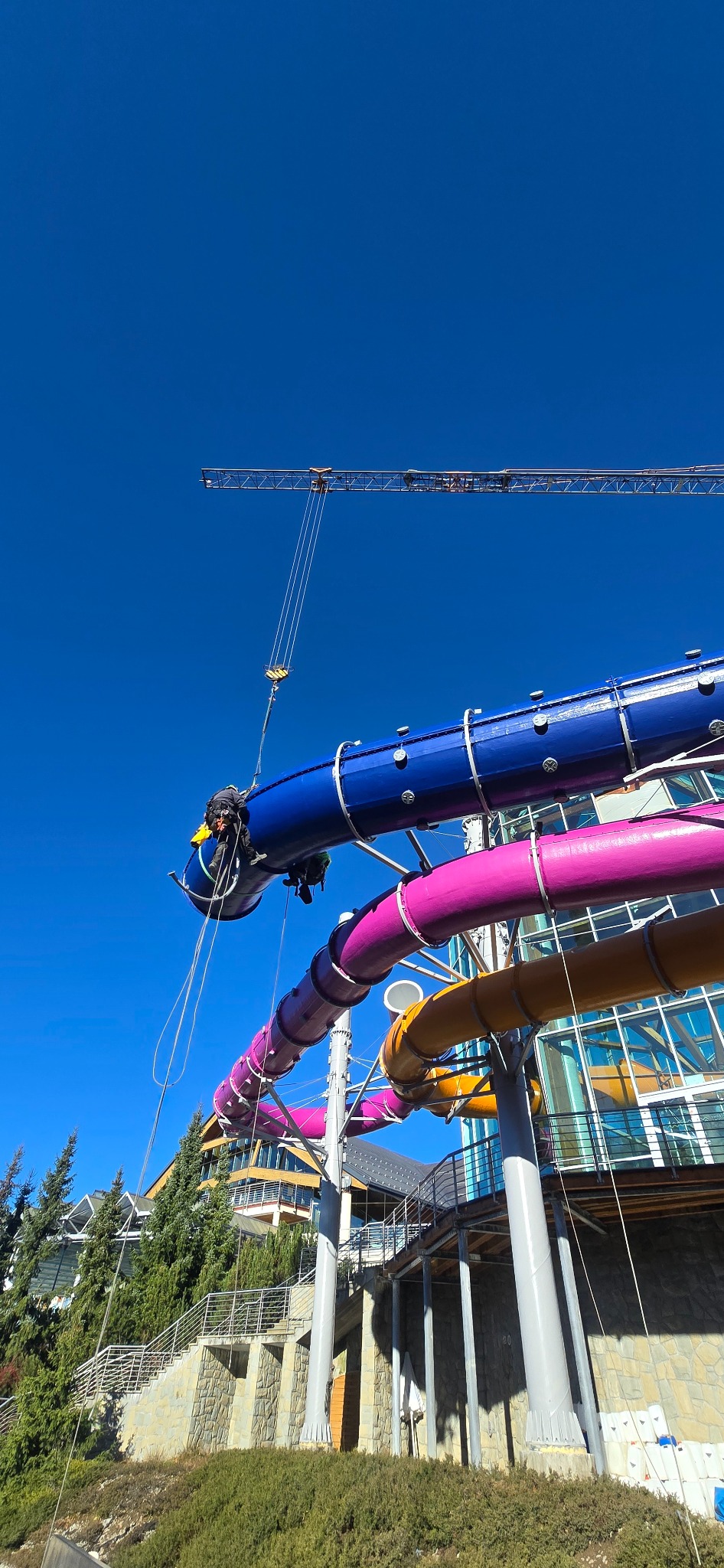 Pracownik zabezpieczony linami, wykonuje prace na wysokości na kolorowej konstrukcji aquaparku. W tle błękitne niebo i żuraw budowlany.