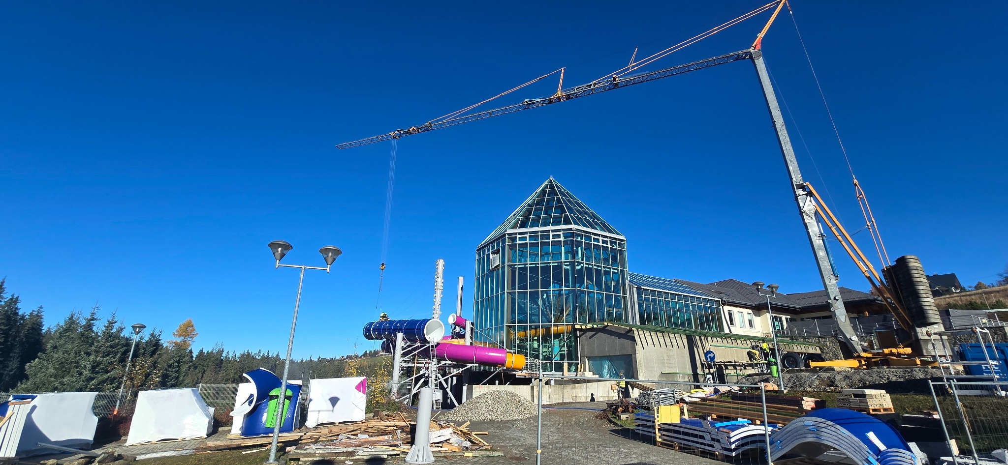 Montaż konstrukcji aquaparku w Warszawie z użyciem dźwigu. Widoczne elementy zjeżdżalni i szklana konstrukcja budynku w słoneczny dzień.