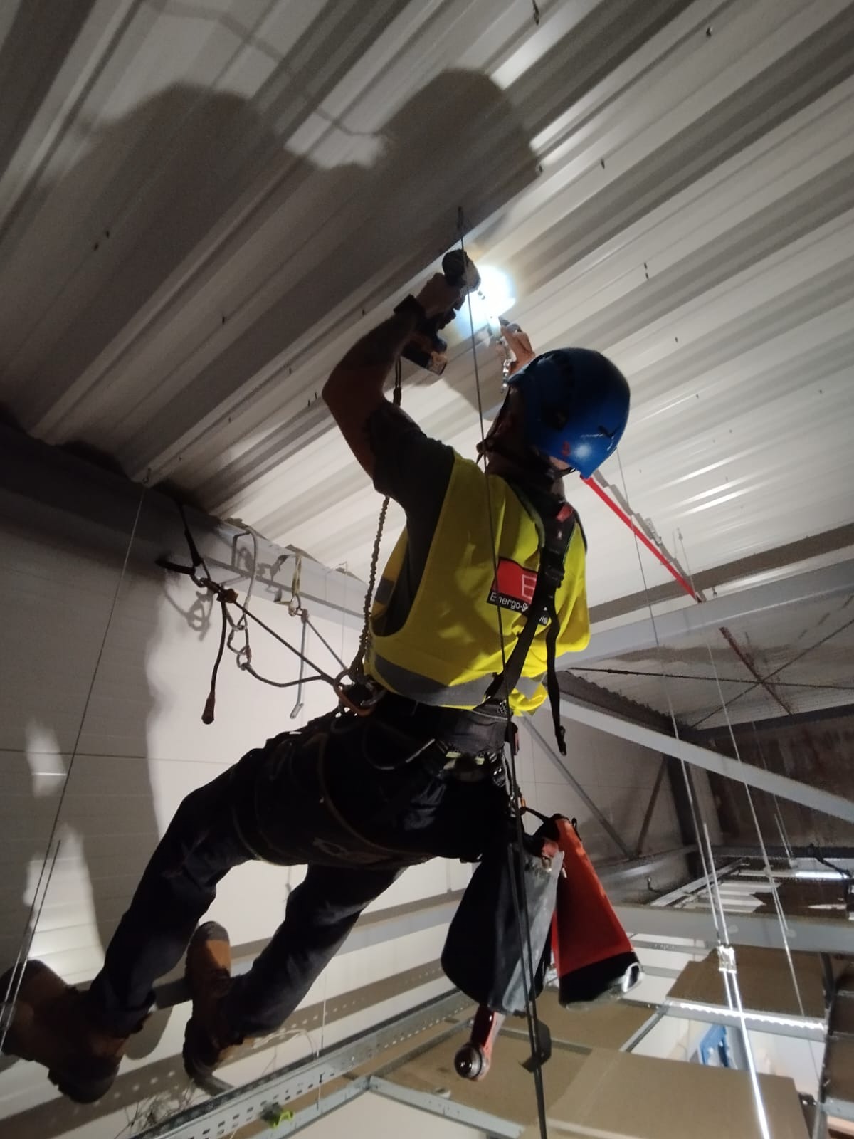 Pracownik w uprzęży, w kasku, wierci w suficie hali. Widoczne liny, narzędzia i oświetlenie. Ujęcie z dołu, podkreślające wysokość i specyfikę pracy.