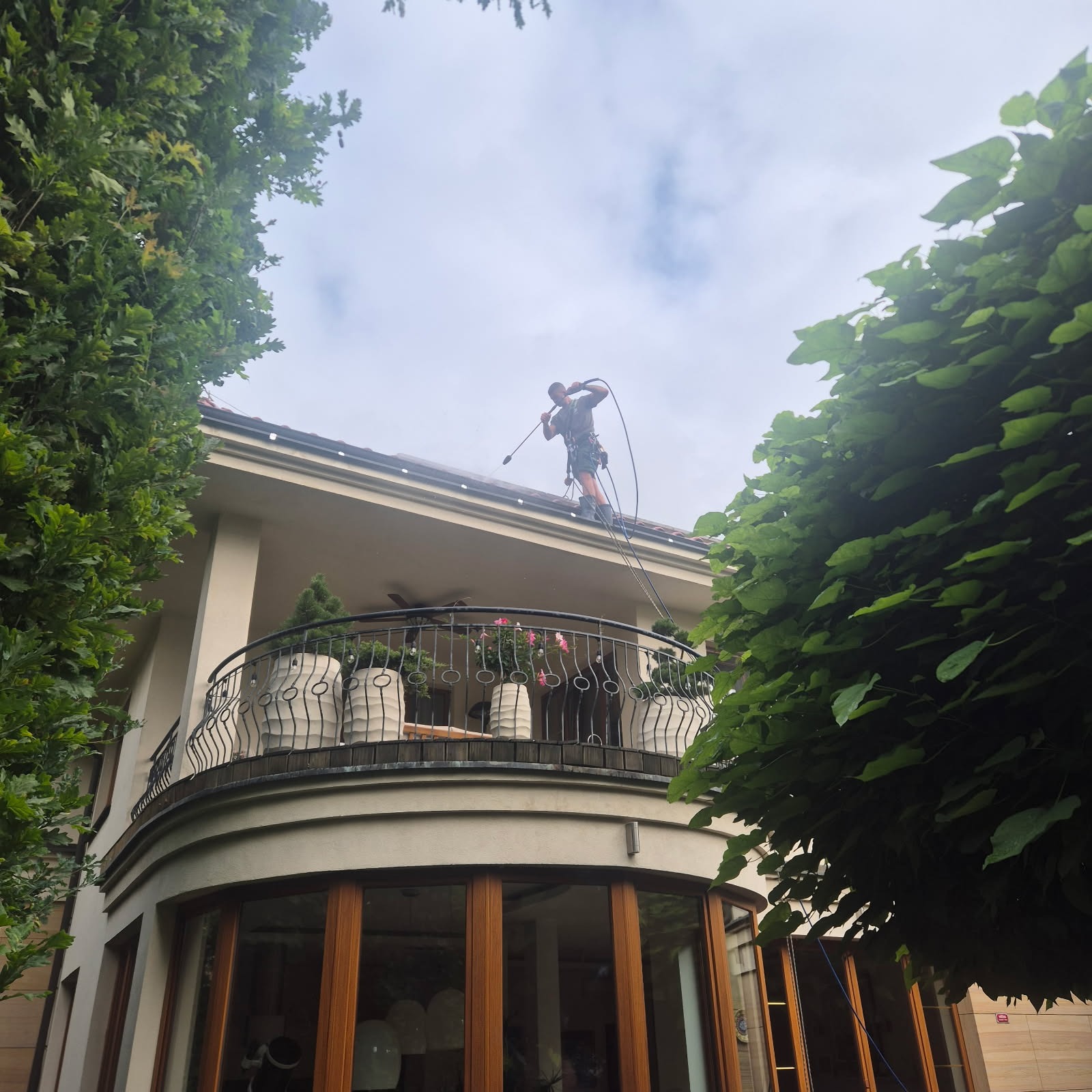 Pracownik w uprzęży czyści dach domu z balkonu za pomocą myjki ciśnieniowej. Widok z dołu, zieleń drzew w kadrze.