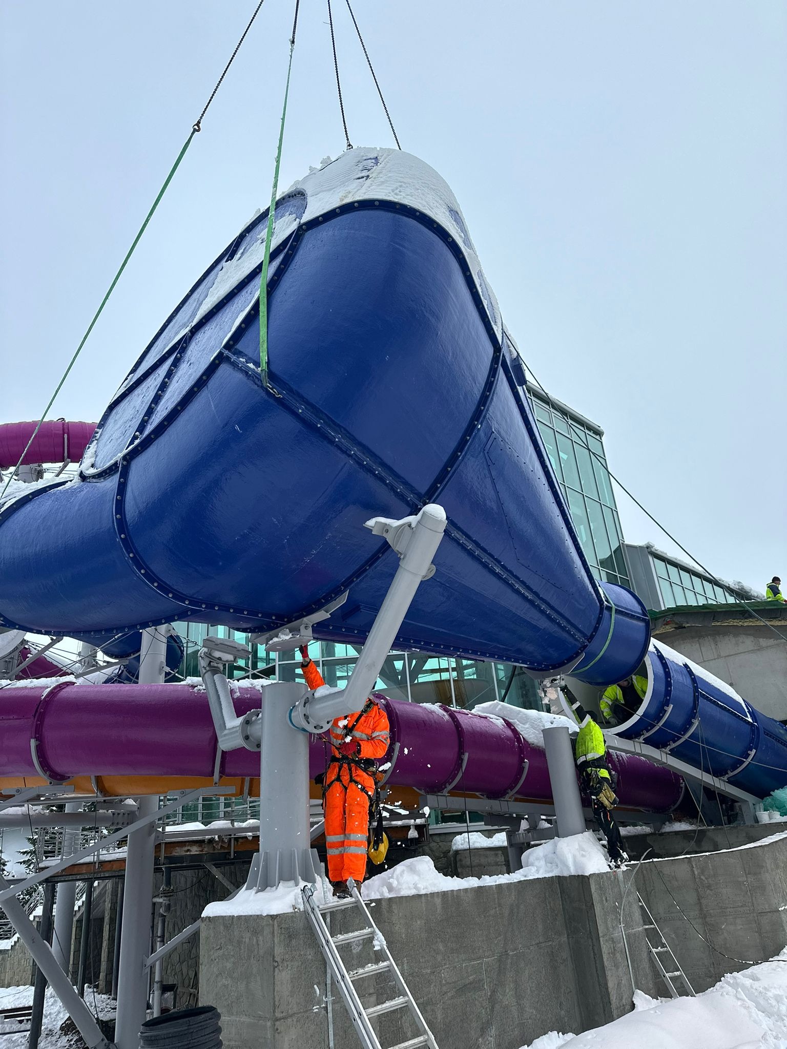 Montaż niebieskiej zjeżdżalni wodnej z elementami fioletowymi. Pracownicy w odzieży ochronnej na wysokości. Zimowa sceneria z widocznym śniegiem.