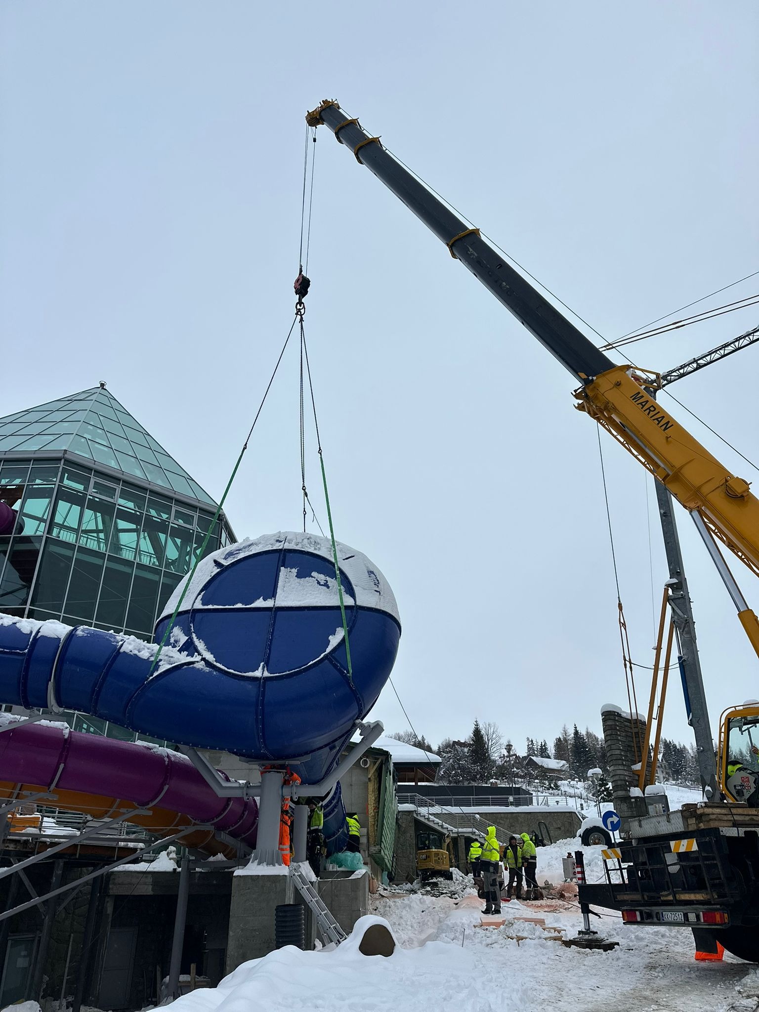 Montaż elementu zjeżdżalni wodnej dźwigiem na zewnątrz budynku w zimowej scenerii. Pracownicy w odblaskowych ubraniach nadzorują proces.