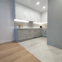 Jasna kuchnia z szarymi szafkami i drewnianą podłogą przechodzącą w płytki imitujące marmur, oświetlenie podszafkowe, czajnik na blacie.