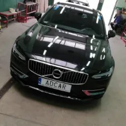 Czarny samochód Volvo S90 z polską tablicą rejestracyjną ADCAR w warsztacie, widoczne odbicia światła na karoserii i niebieska taśma na przedniej szybie.