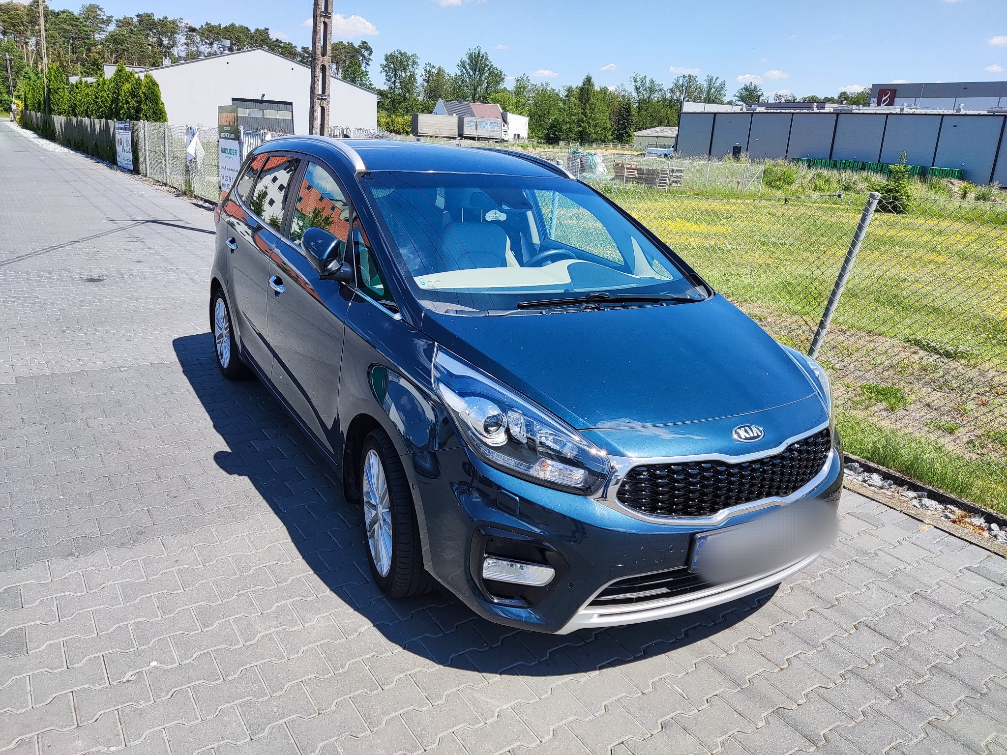 Błyszczący, granatowy samochód Kia Carens zaparkowany na brukowanej powierzchni w słoneczny dzień, z widocznym logo marki na grillu i srebrnymi relingami dachowymi.