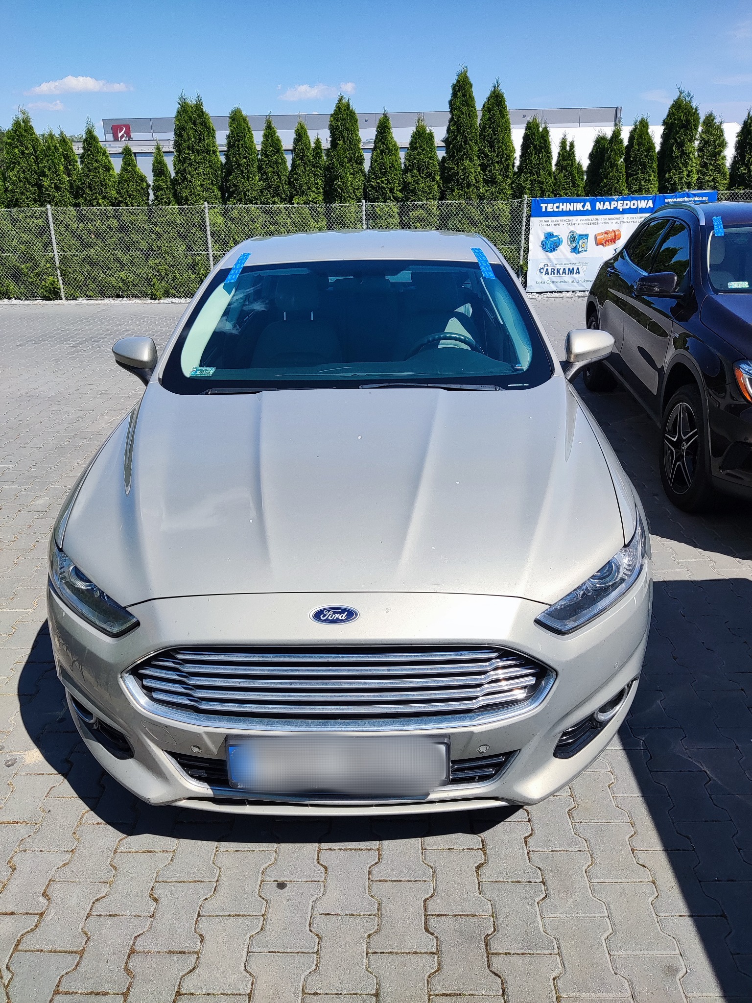 Przód srebrnego Forda Fusion na parkingu, z niebieską taśmą zabezpieczającą na przedniej szybie, obok czarnego SUV-a. W tle widoczny budynek z zielonymi tujami i baner reklamowy.