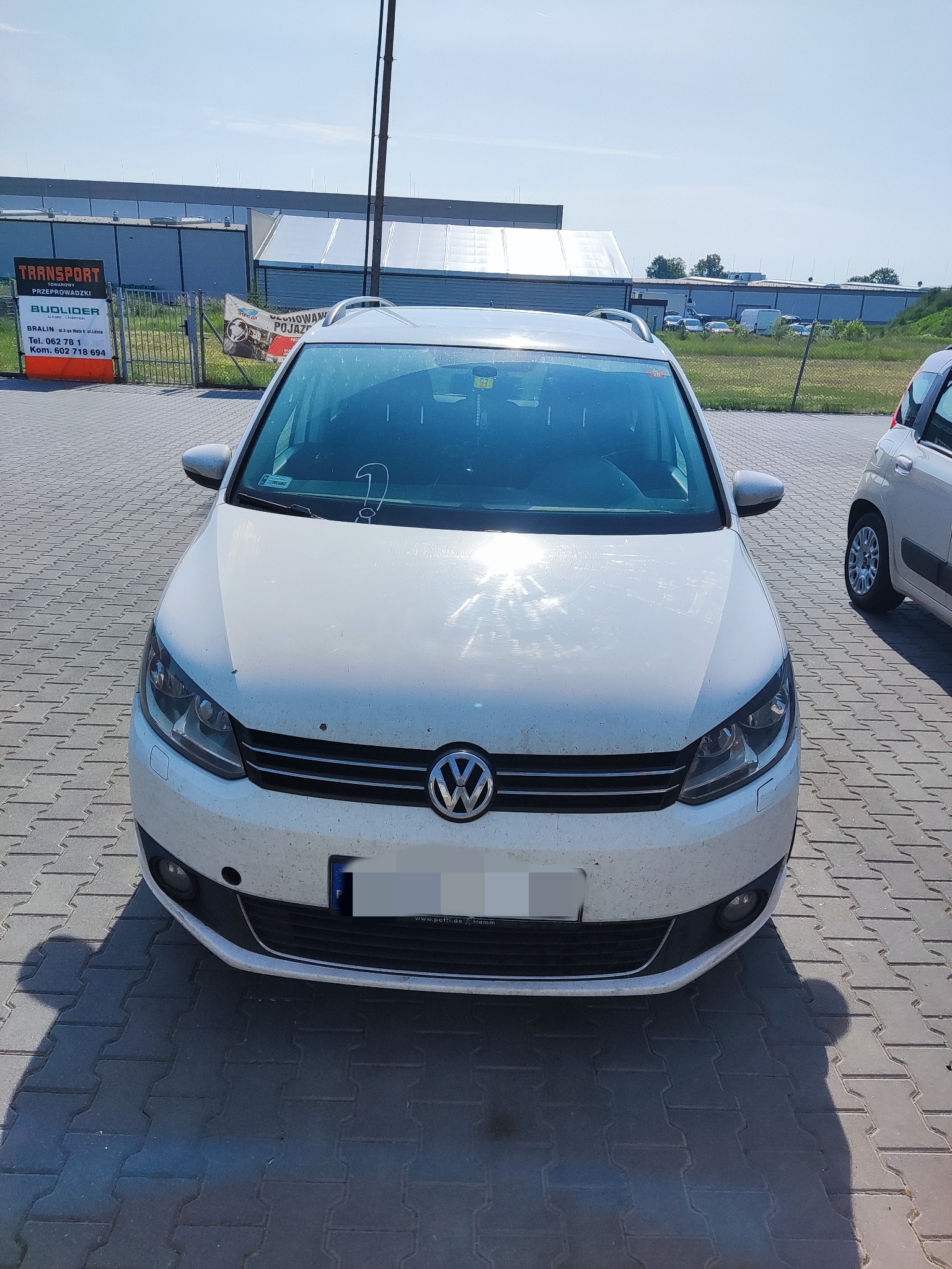 Przód białego, lekko zabrudzonego kombi Volkswagen na brukowanym parkingu w słoneczny dzień. W tle budynek magazynowy i ogrodzenie z reklamą firmy transportowej.