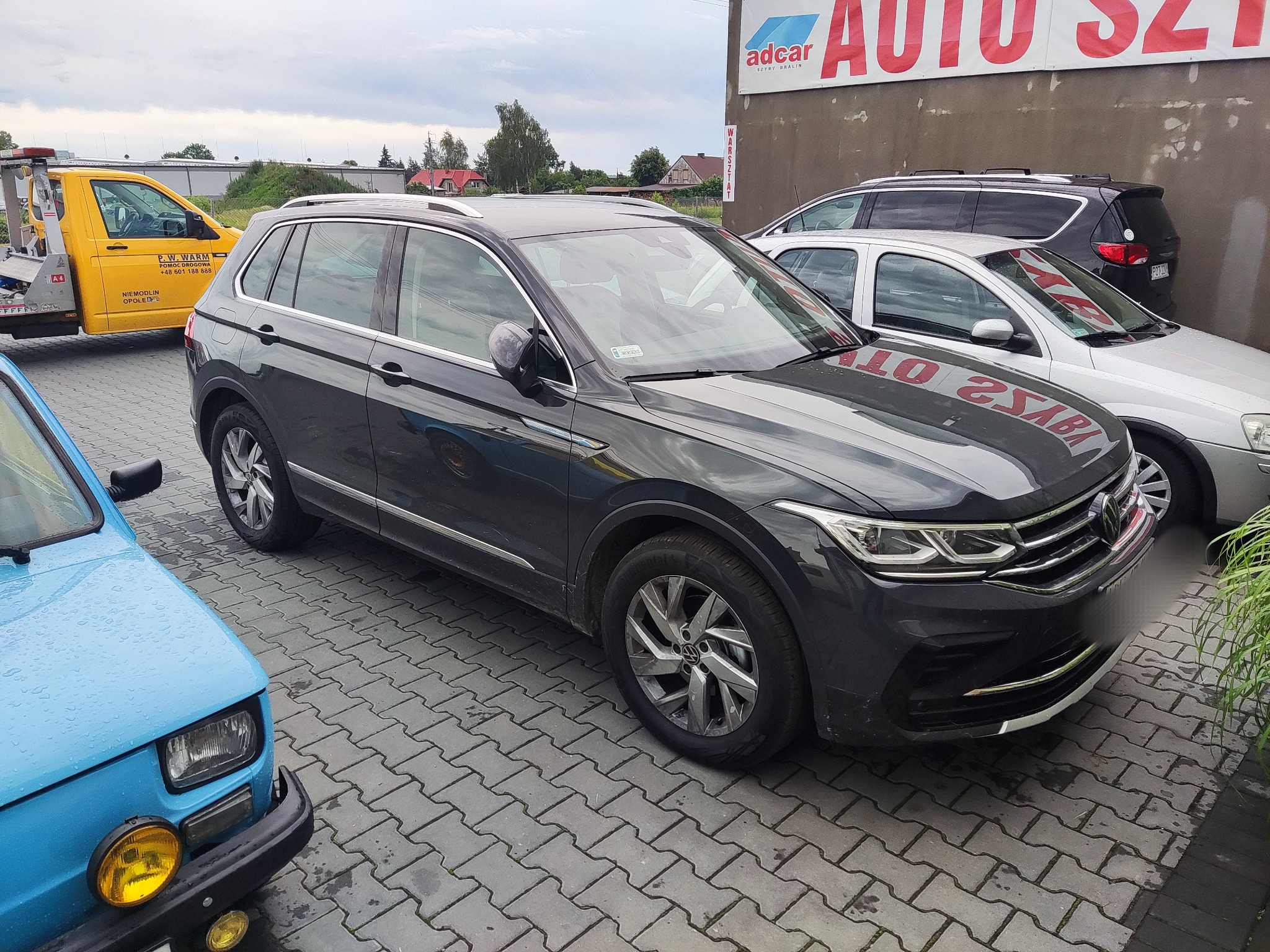 Ciemnoszary Volkswagen Tiguan zaparkowany na brukowanym placu przed budynkiem z szyldem warsztatu samochodowego, obok stoi żółty holownik pomocy drogowej i niebieski Fiat 126p.