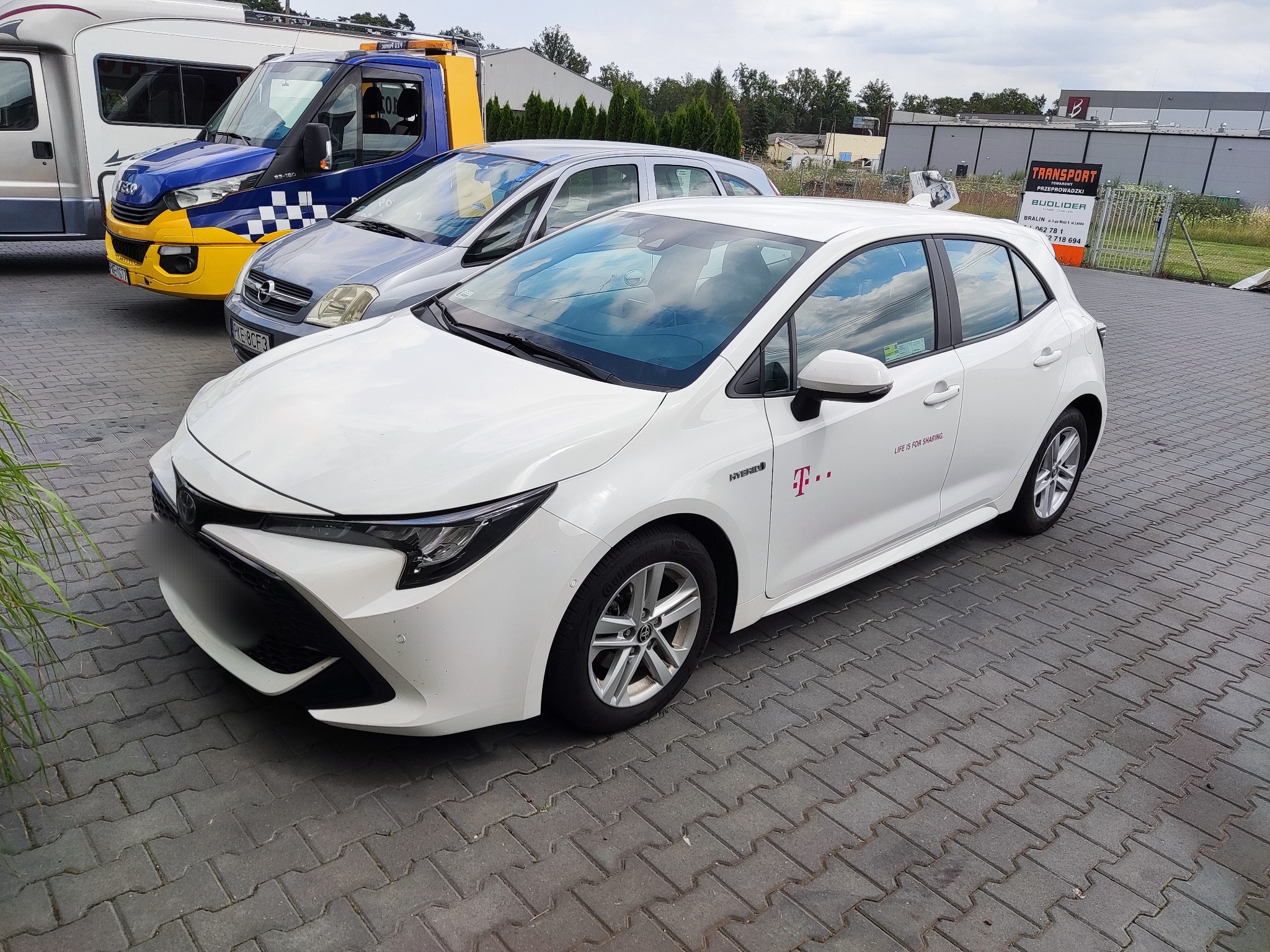 Biała Toyota Corolla Hybrid z logo T-Mobile parkująca na brukowanym placu, w tle laweta i szary samochód osobowy.