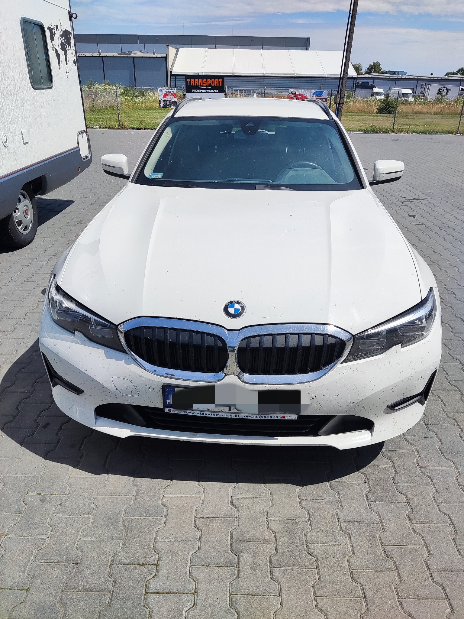 Białe BMW serii 3 kombi, lekko zabrudzone, zaparkowane na kostce brukowej, widok z przodu, obok zaparkowany kamper, w tle budynek z napisem TRANSPORT.