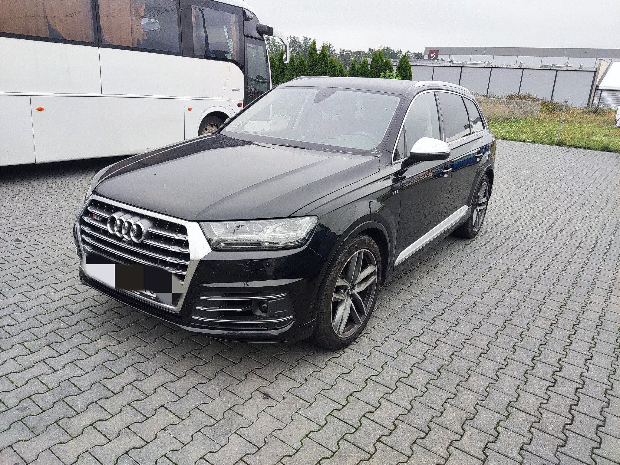 Czarny SUV Audi Q7 zaparkowany na szarej kostce brukowej, z widocznym autobusem w tle i budynkiem przemysłowym w oddali.