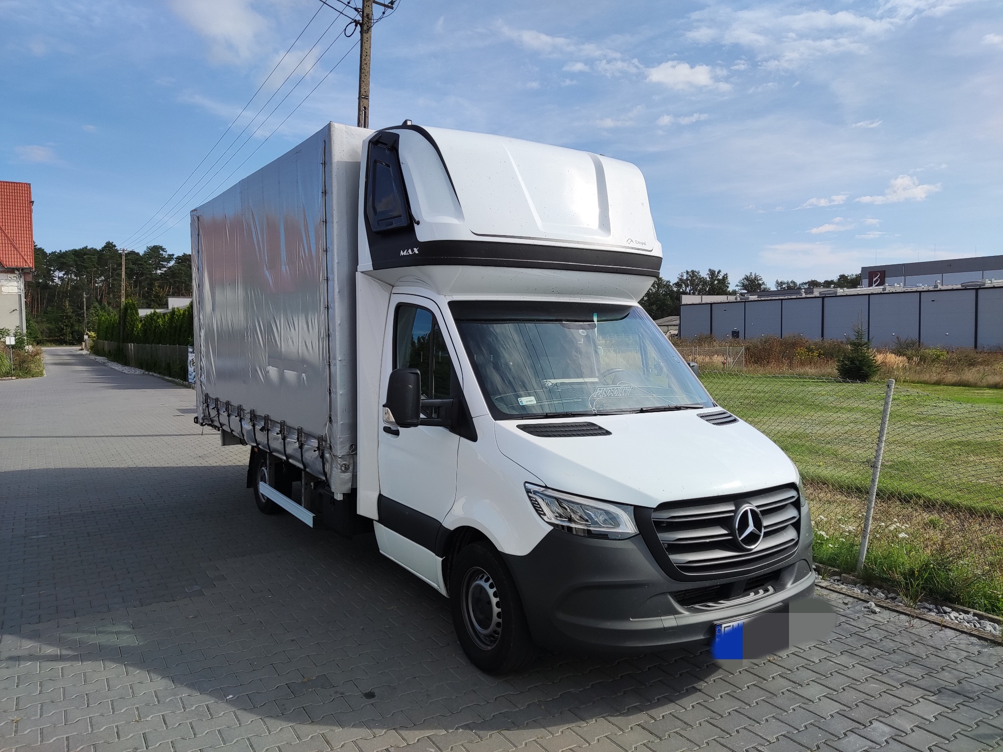 Biały samochód dostawczy Mercedes-Benz Sprinter z plandeką na parkingu z kostki brukowej w słoneczny dzień.