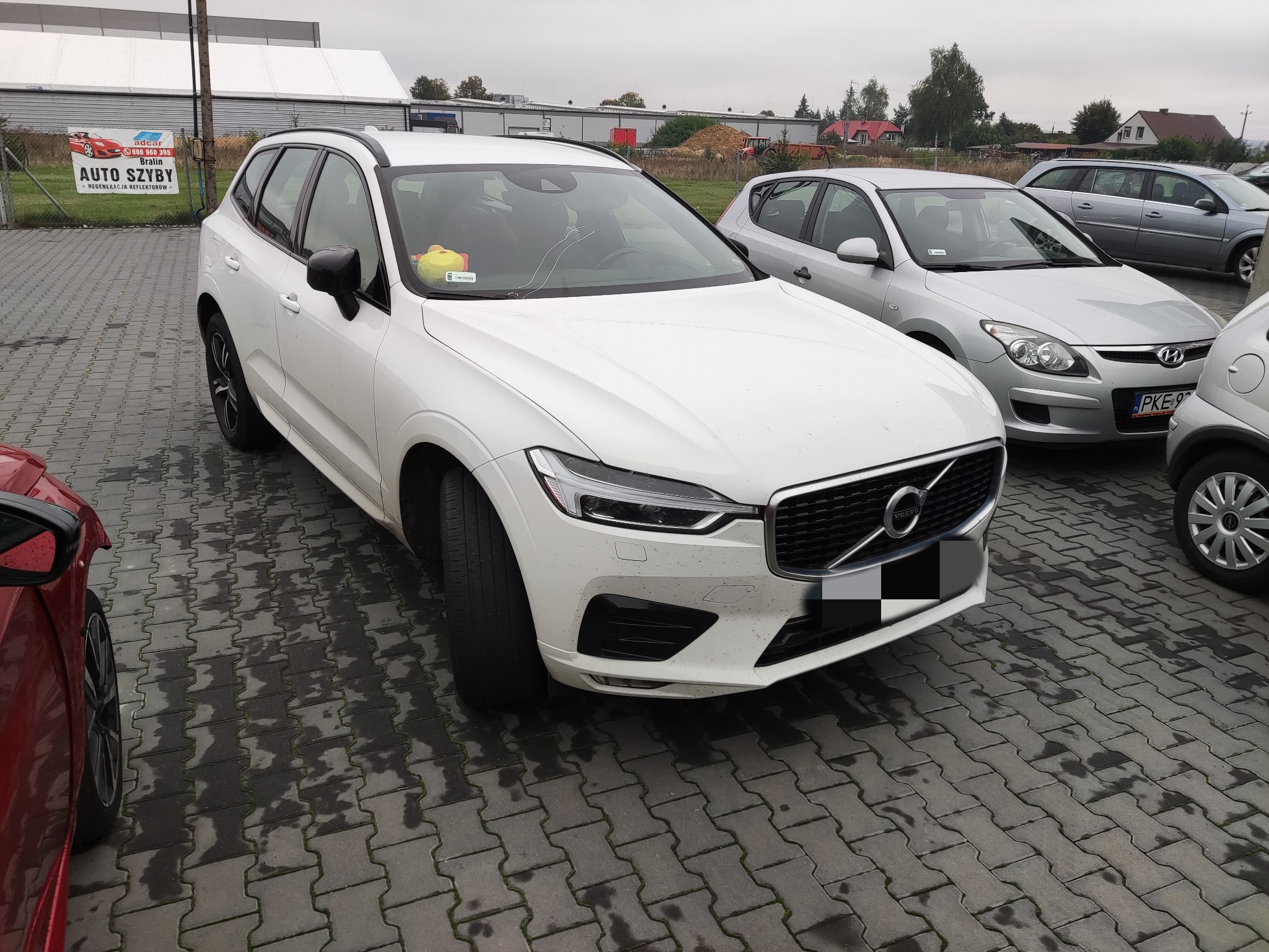 Białe Volvo XC60 zaparkowane na mokrej kostce brukowej, widok z przodu, na przedniej szybie żółta maskotka.