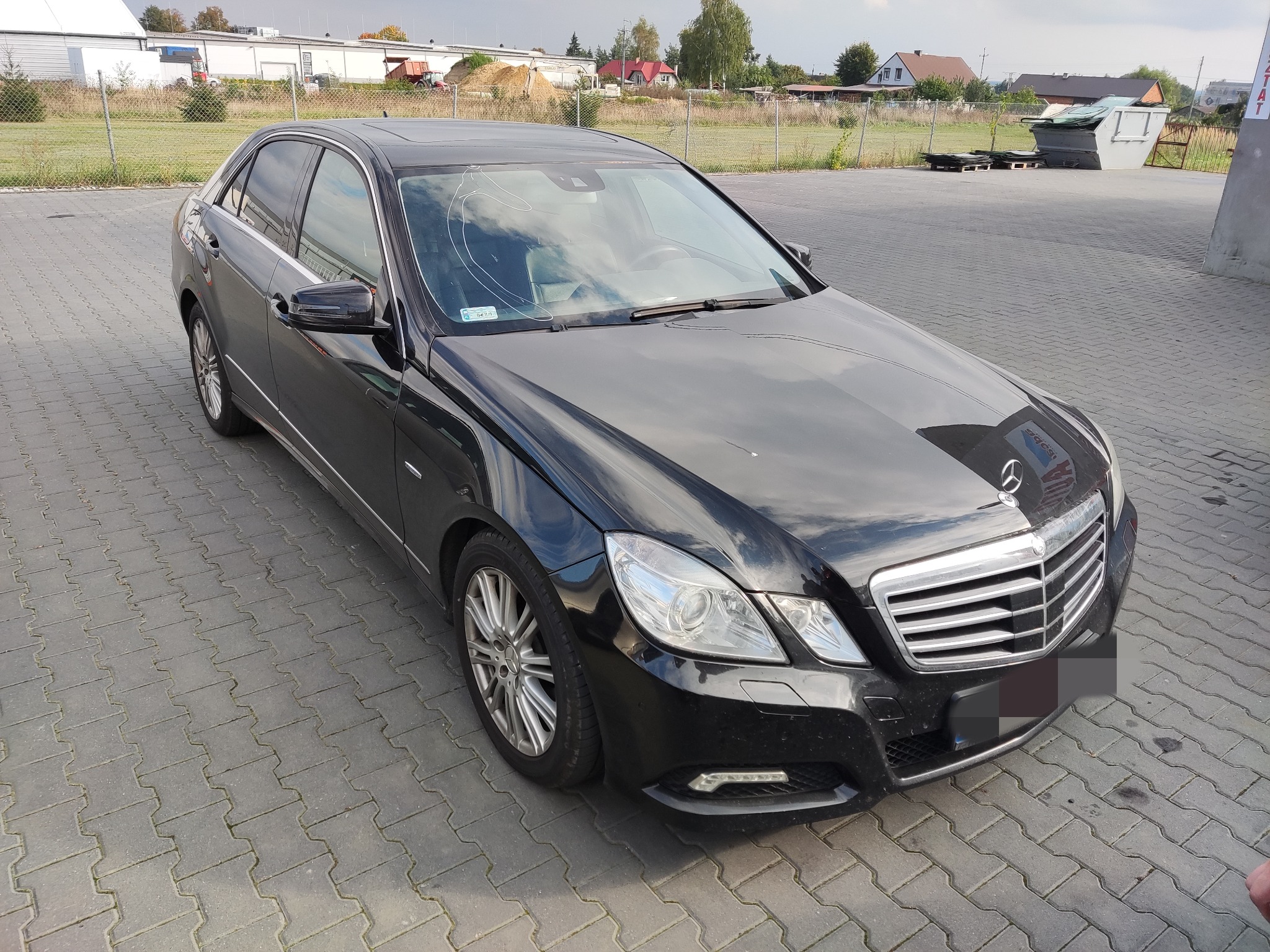 Czarny samochód osobowy marki Mercedes z pękniętą przednią szybą, zaparkowany na betonowej kostce brukowej. W tle widoczne zabudowania gospodarcze i ogrodzenie z siatki.