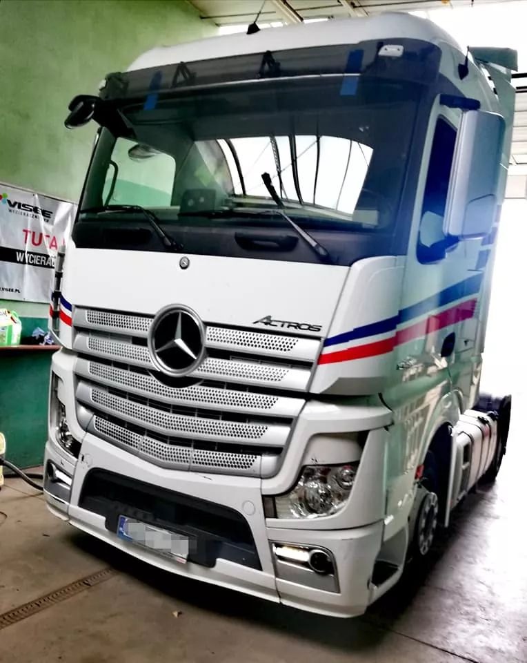 Biała ciężarówka Mercedes Actros z niebiesko-czerwonymi pasami, zaparkowana w warsztacie z widoczną reklamą środków czyszczących w tle.