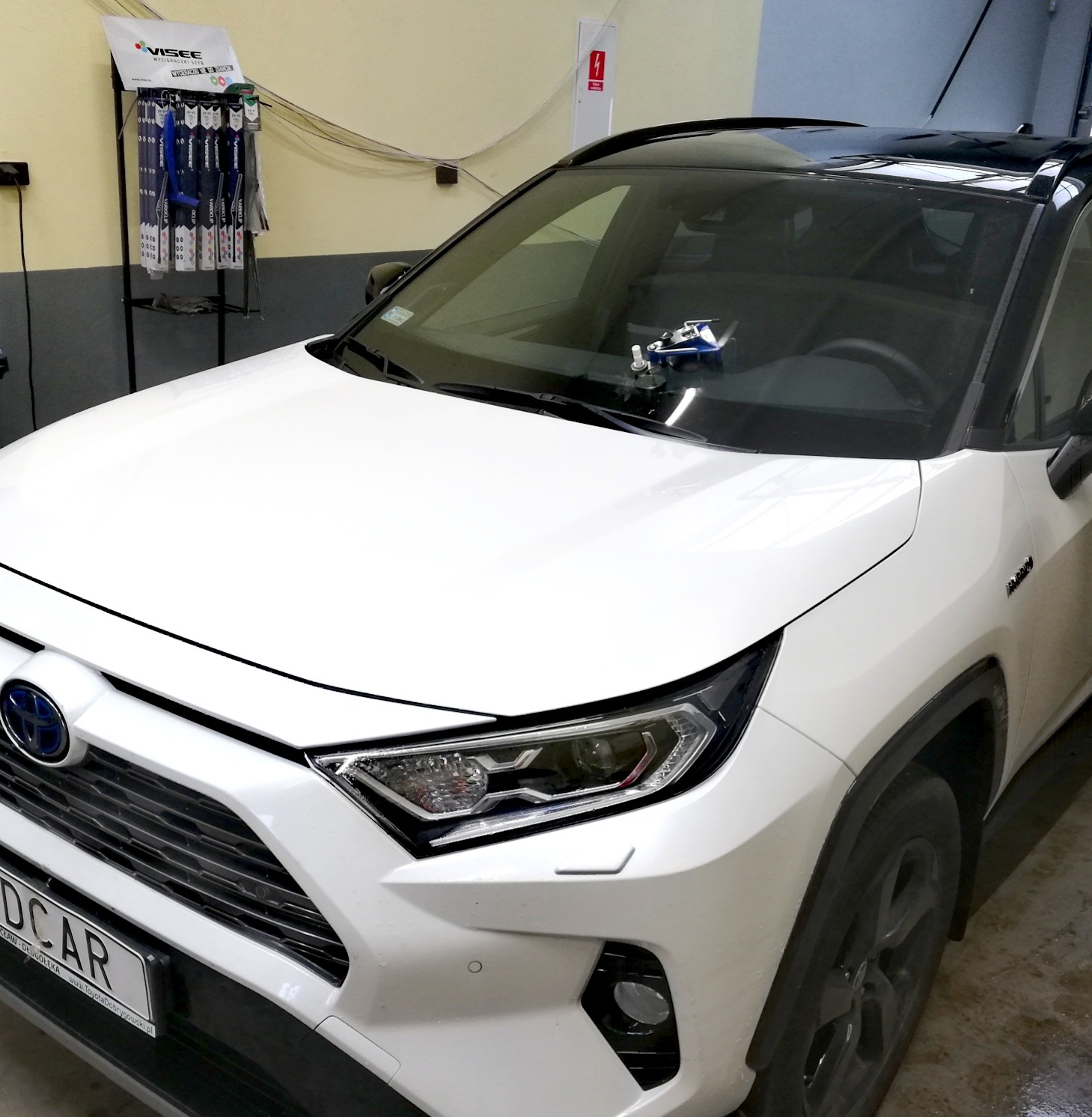 Biała Toyota RAV4 z urządzeniem do naprawy odprysków na przedniej szybie, widoczne akcesoria do wycieraczek na półce w tle.