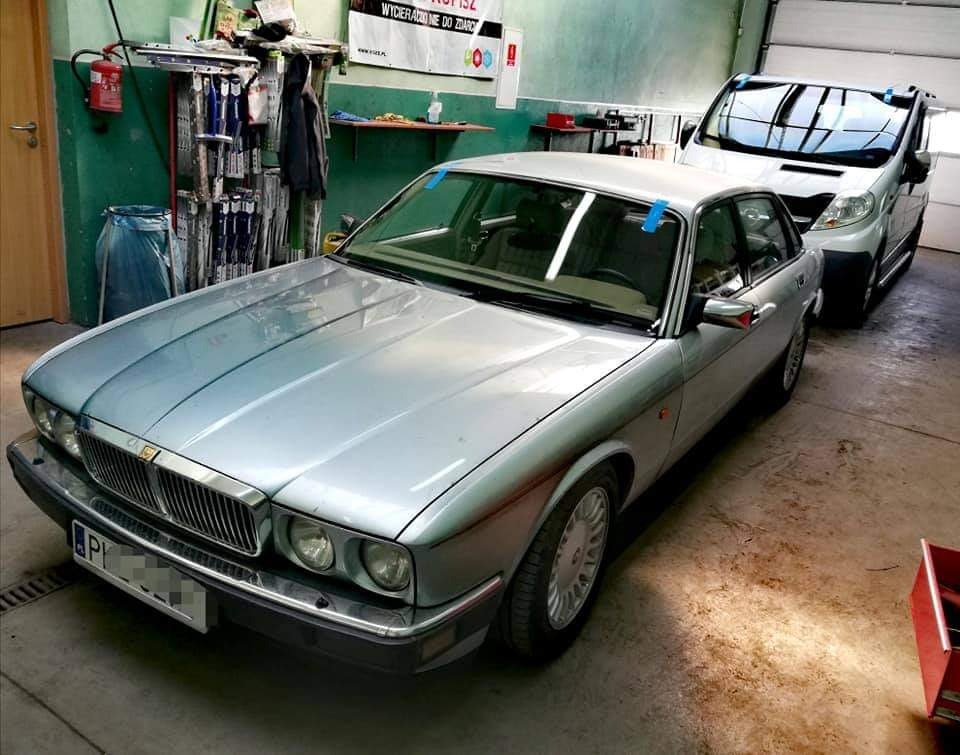 Srebrny Jaguar XJ w warsztacie, z niebieską taśmą zabezpieczającą, obok zaparkowany biały van z folią na przedniej szybie, narzędzia i akcesoria warsztatowe w tle.