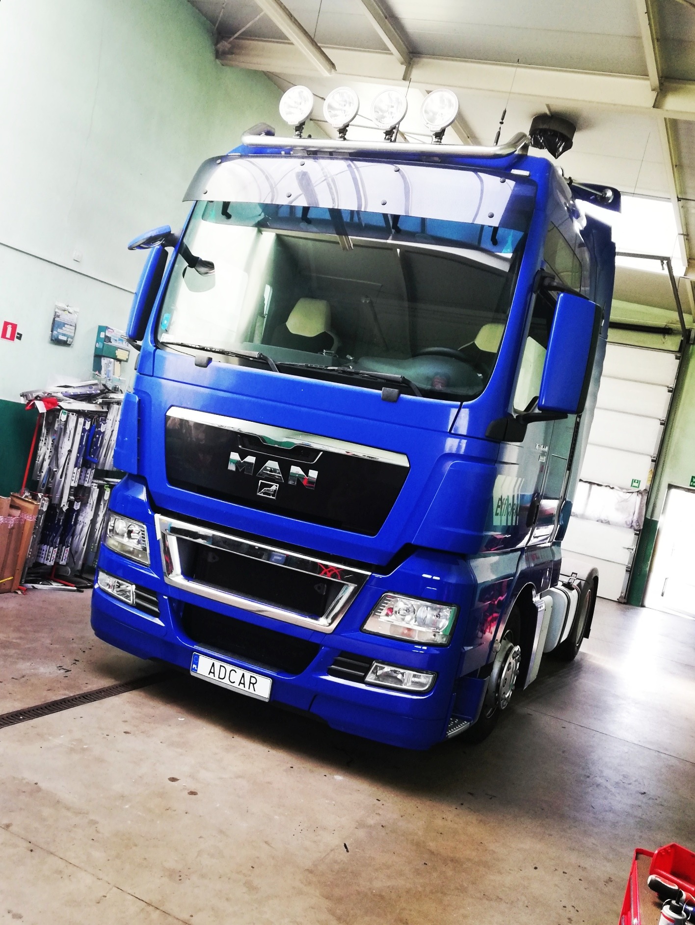 Niebieski ciągnik siodłowy marki MAN z dodatkowymi reflektorami na dachu, zaparkowany w warsztacie z widocznym logo firmy na tablicy rejestracyjnej.