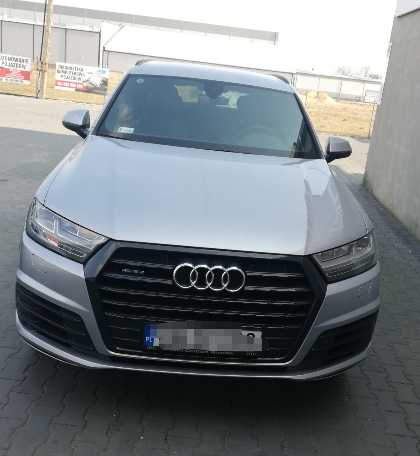 Srebrny SUV marki Audi Q7 z widocznym logo producenta na przedniej atrapie, zaparkowany na kostce brukowej przed budynkiem z reklamą diagnostyki komputerowej pojazdów w tle.