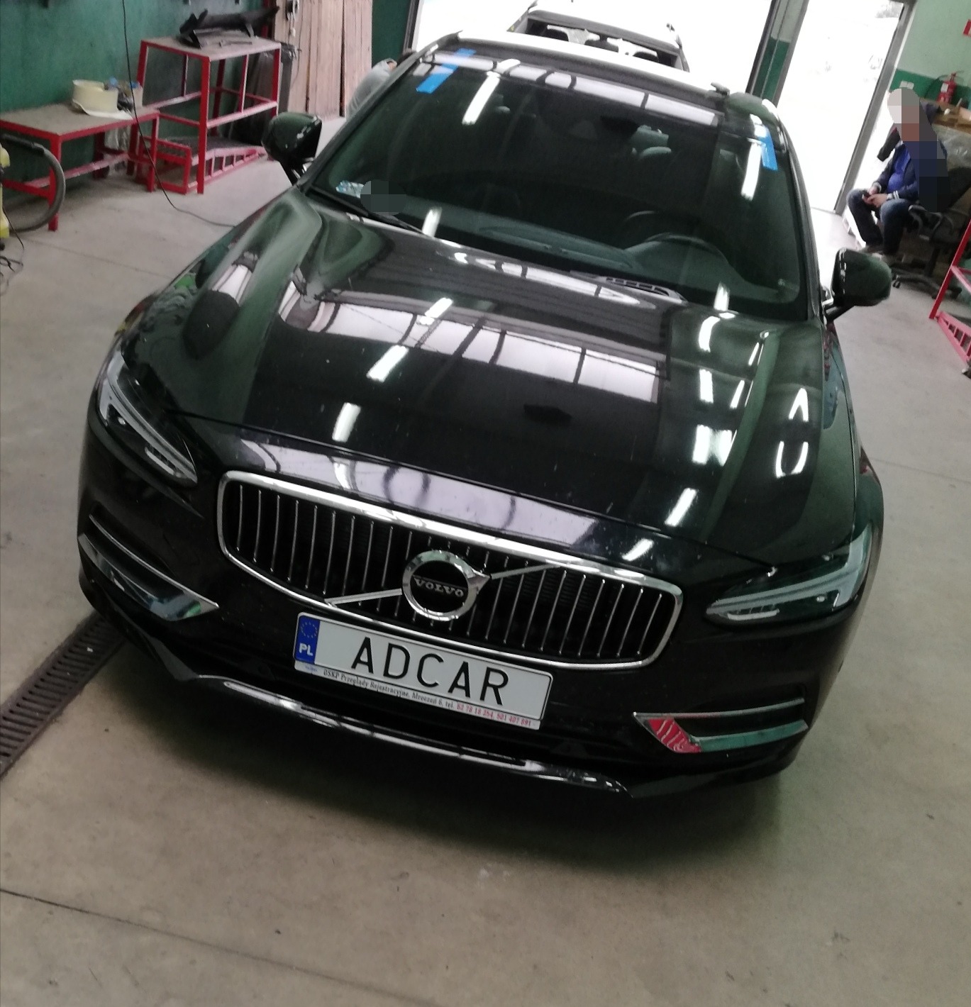 Czarny samochód Volvo S90 z polską tablicą rejestracyjną ADCAR w warsztacie, widoczne odbicia światła na karoserii i niebieska taśma na przedniej szybie.