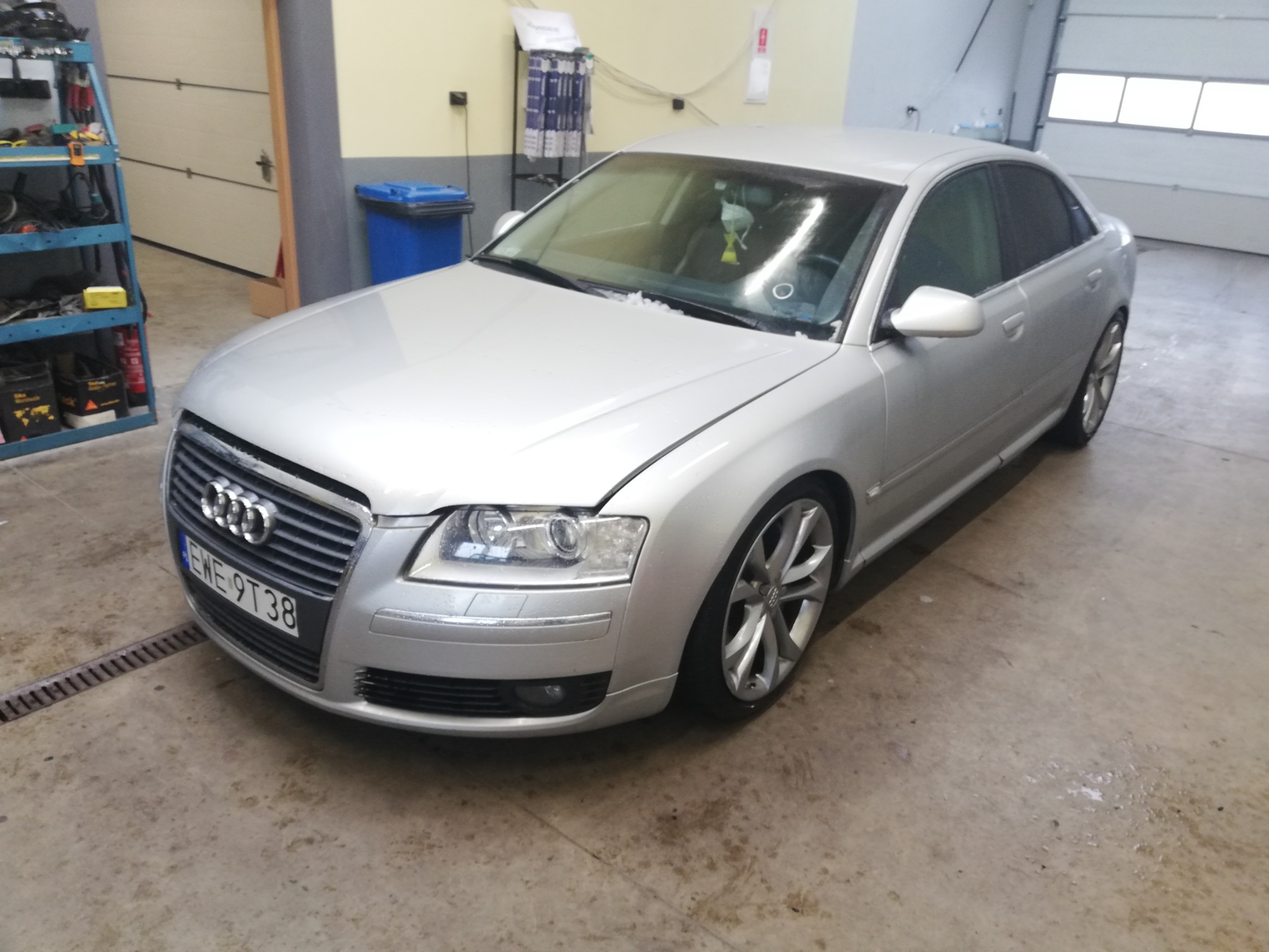 Srebrny samochód Audi A8 z otwartą maską, zaparkowany w warsztacie samochodowym z widocznymi narzędziami i akcesoriami na półkach w tle