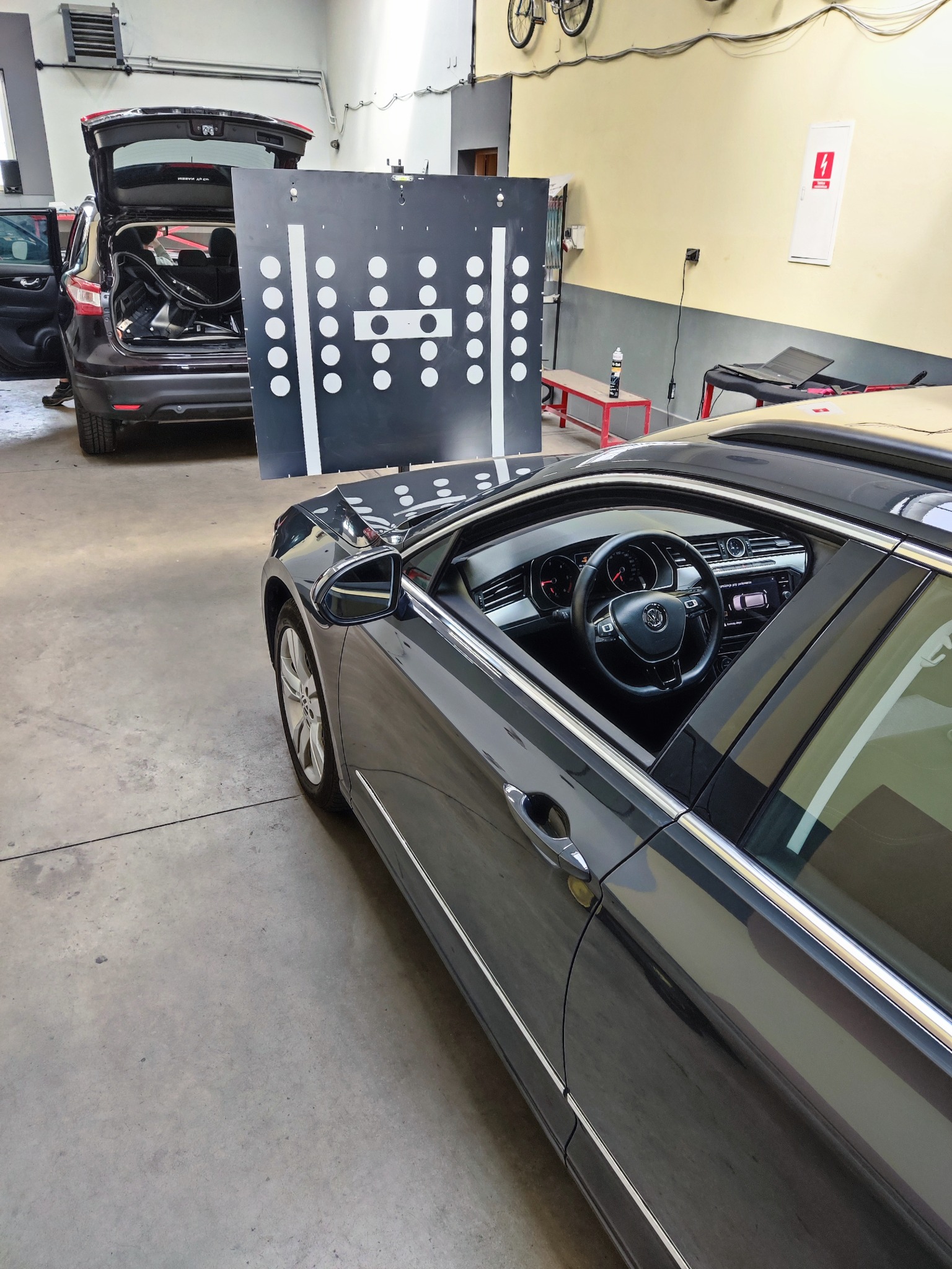 Szary samochód osobowy w warsztacie podczas ustawiania geometrii kół. Widoczna tablica kalibracyjna z białymi punktami i otwarty bagażnik innego pojazdu w tle.