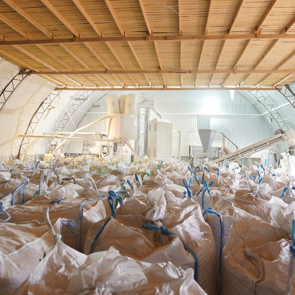 Magazyn wypełniony workami z gotowym pelletem, w tle linia produkcyjna w hali o konstrukcji łukowej.