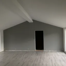 Wnętrze pokoju z szarą ścianą, jasnoszarą podłogą z paneli i białym sufitem z widoczną belką konstrukcyjną, duże okna z białymi ramami po bokach, widoczne puszki elektryczne na ścianie, otwór...