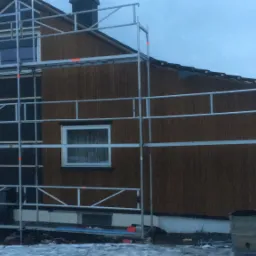 Dom w trakcie budowy, widoczne rusztowanie, częściowo ocieplona ściana i drewniana elewacja, zimowa sceneria.