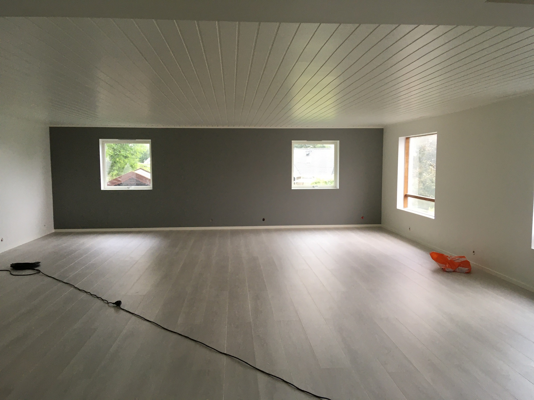 Puste, jasne wnętrze po remoncie: świeżo ułożone panele podłogowe w odcieniach szarości, pomalowane ściany w bieli i szarości, biały sufit z boazerią, dwa okna z widokiem na zieleń, kabel...