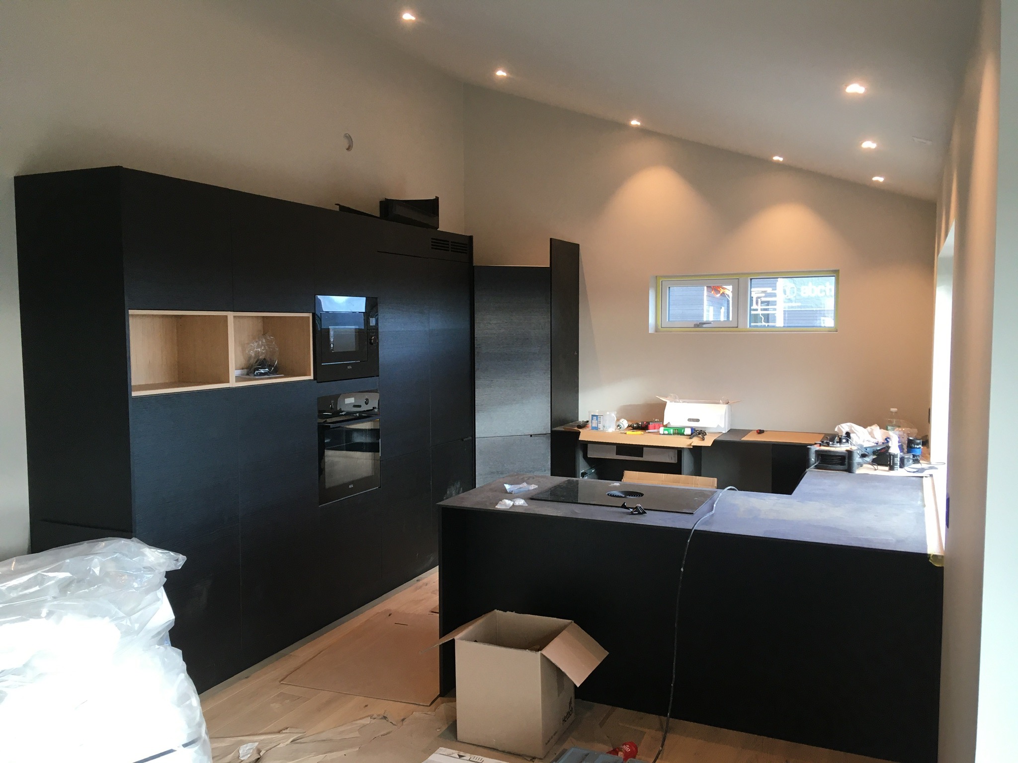 Montaż nowoczesnej, czarnej zabudowy kuchennej z wbudowanym piekarnikiem i płytą grzewczą, widoczne kartony i folia ochronna na podłodze, surowe oświetlenie punktowe.