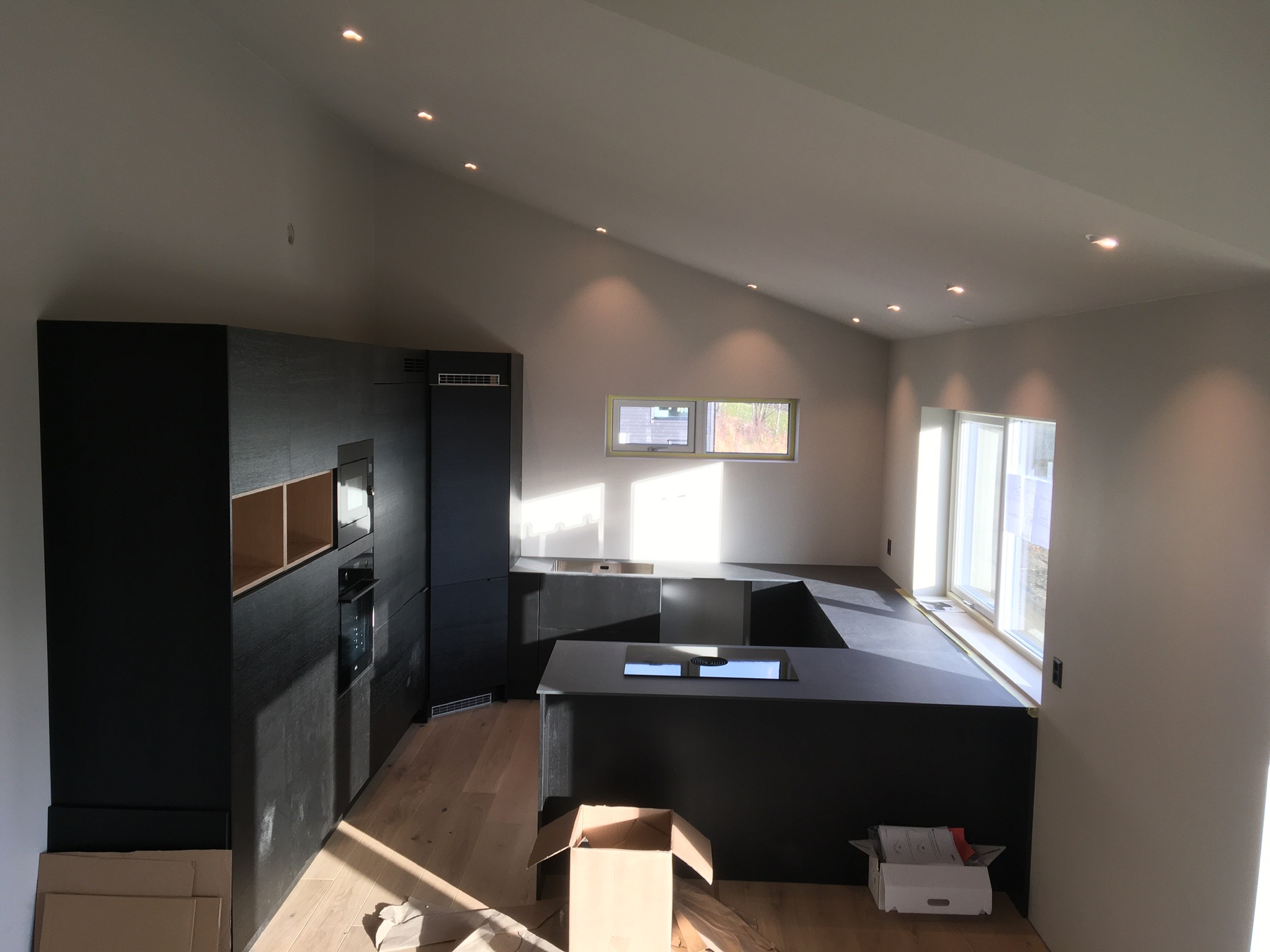 Wnętrze kuchni w trakcie prac wykończeniowych, widoczne czarne szafki z drewnianą fakturą, drewniana podłoga, kartony po sprzęcie, sufit ze skośnym profilem i oświetleniem punktowym.