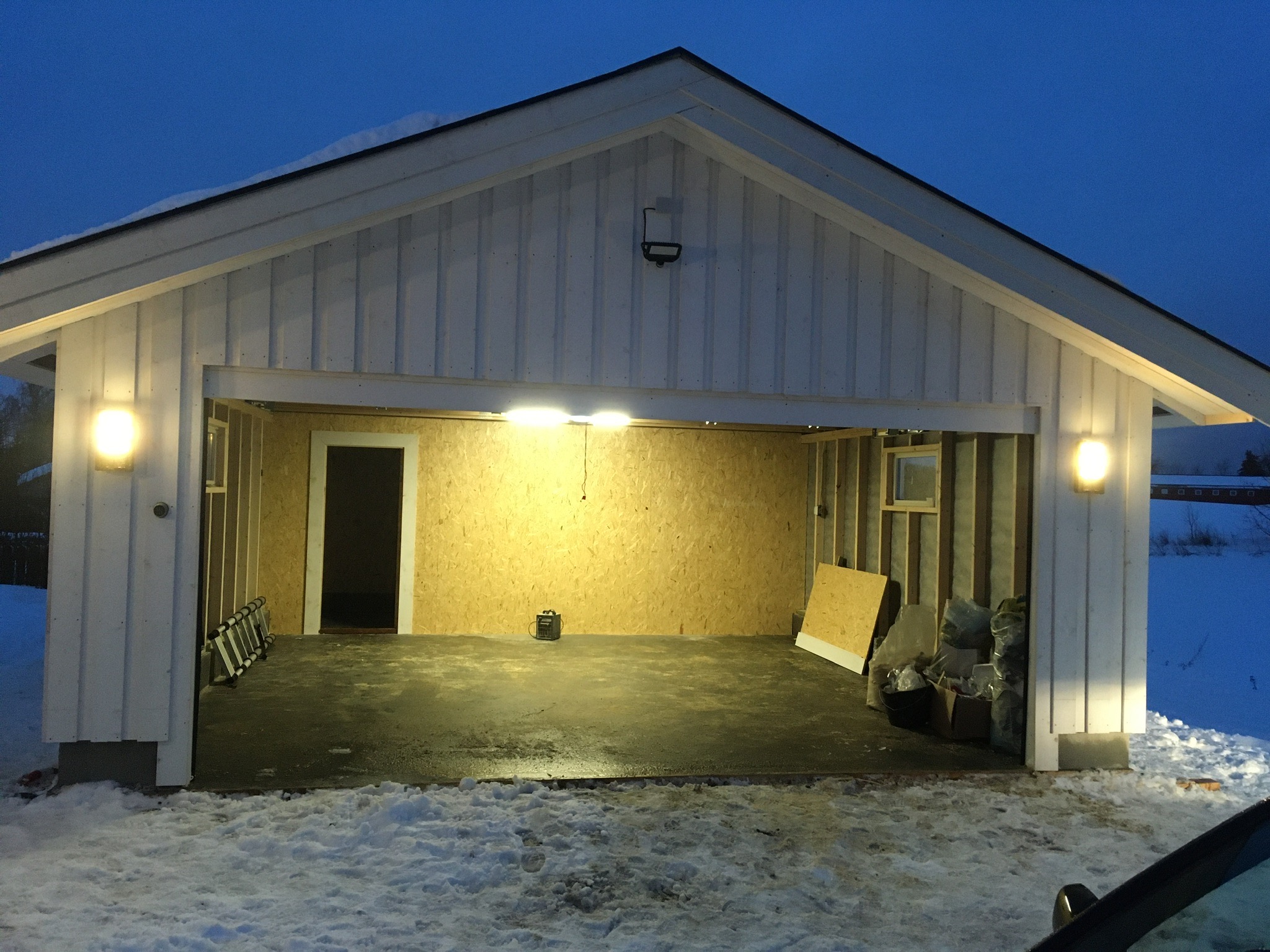 Wnętrze garażu w trakcie budowy domu szkieletowego, widoczne oświetlenie, ściany z płyt OSB, betonowa posadzka, drabina i materiały budowlane, na zewnątrz śnieg.