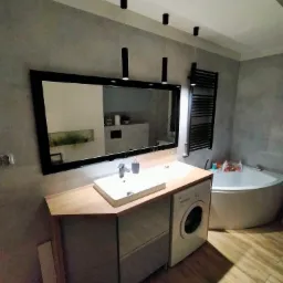 Nowoczesna łazienka z umywalką nablatową, dużym lustrem w czarnej ramie, wiszącymi lampami tubami, grzejnikiem drabinkowym i pralką zabudowaną pod blatem. W tle widoczna wanna.