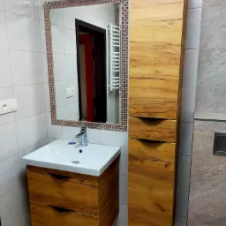 Nowoczesna łazienka z drewnianą szafką pod umywalką i wysoką szafką z szufladami o podobnym wykończeniu, lustro z mozaikową ramą, białe płytki na ścianach.