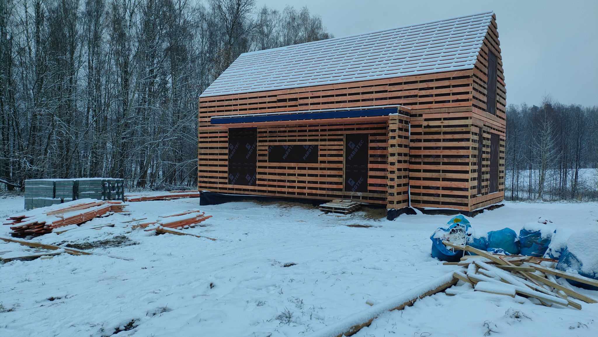 Surowy stan konstrukcji budynku w technologii szkieletowej, widoczne drewniane elementy, folia paroizolacyjna w otworach okiennych i drzwiowych, zasypane śniegiem otoczenie z drzewami w tle.
