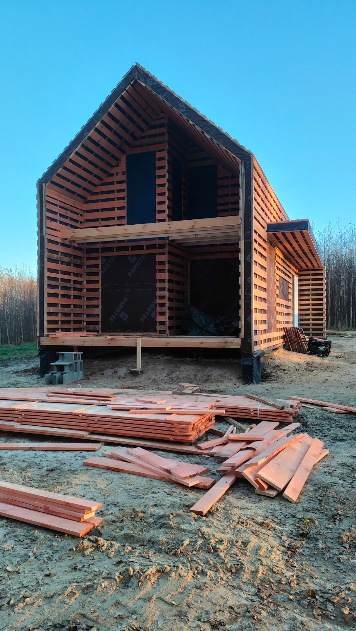Konstrukcja dwukondygnacyjnego domu w trakcie budowy, widoczne drewniane elementy szkieletu i czarna membrana, stos desek na pierwszym planie.